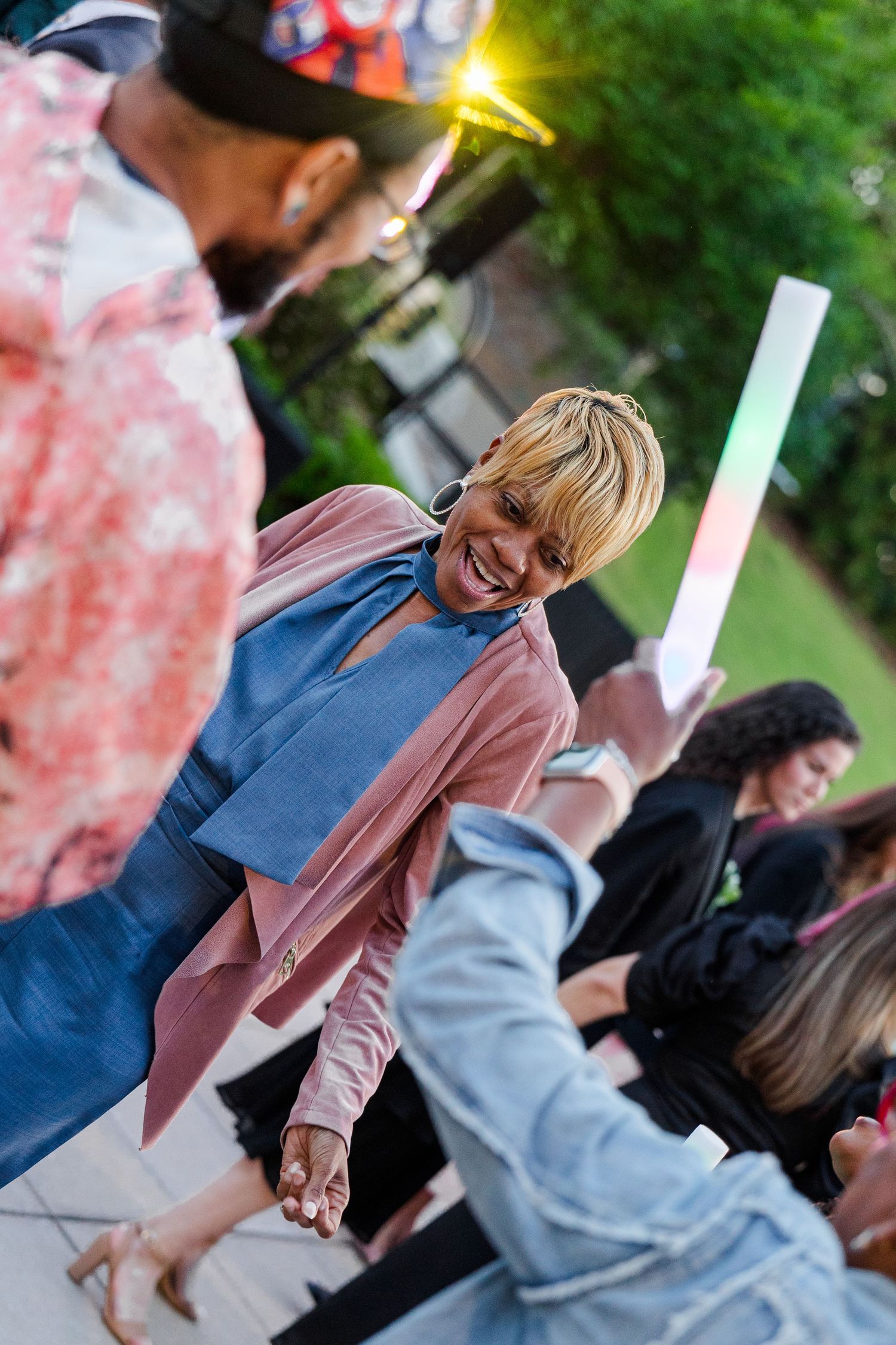 A smiling woman with blonde hair in a pink blazer and denim top greets someone at an outdoor event.