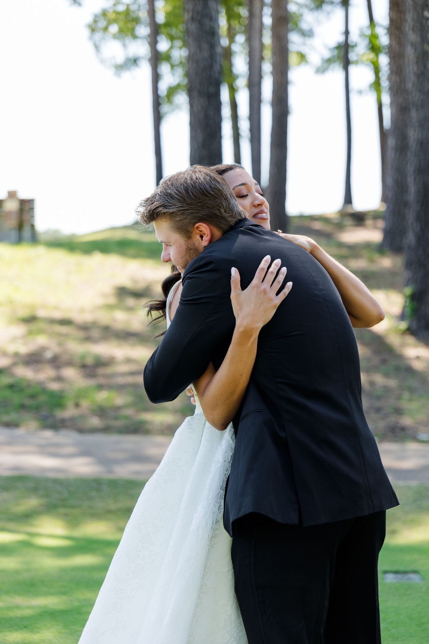 Bride and groom share an emotional first look hug outdoors, surrounded by tall pine trees on a sunny wedding day.