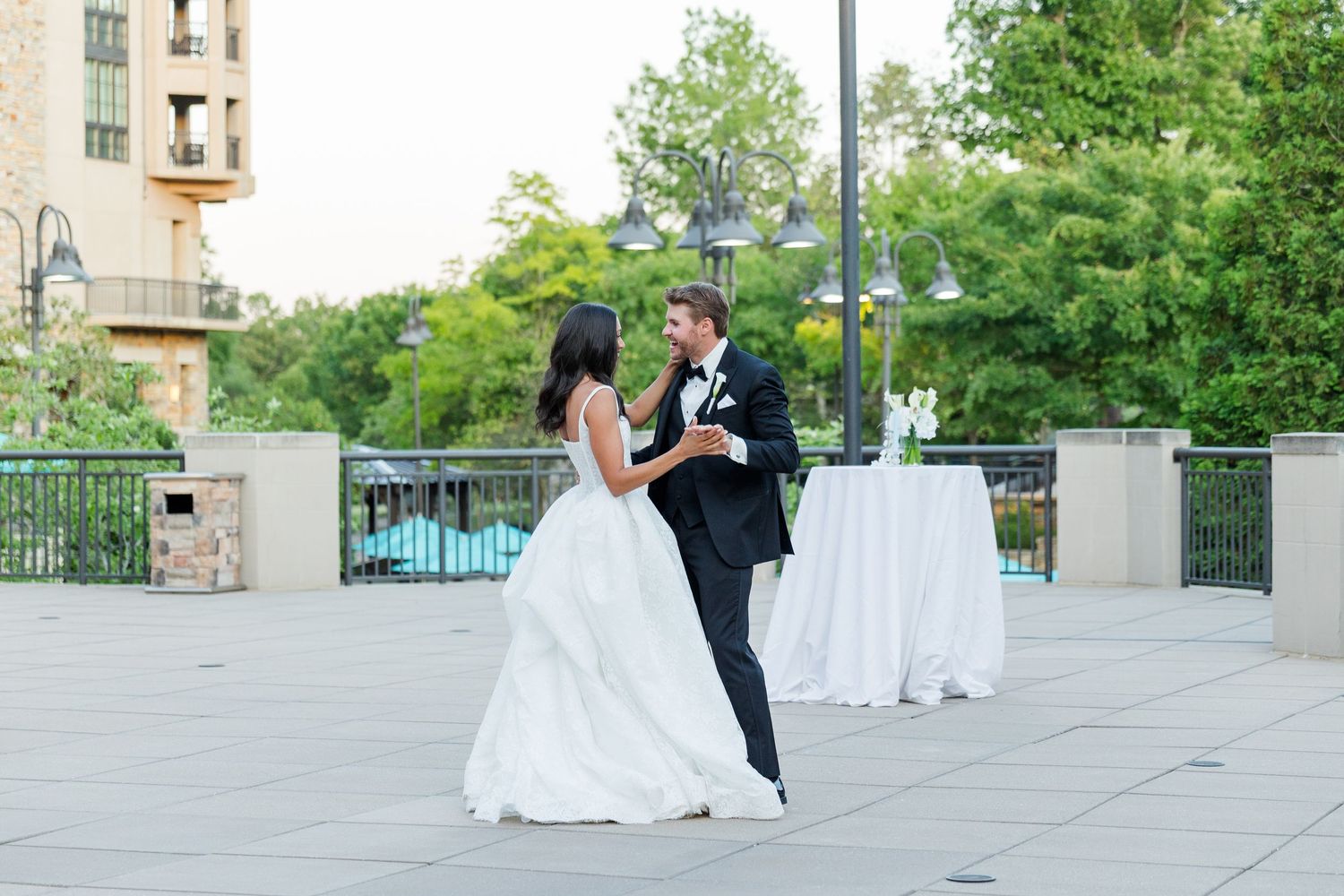 Bride and groom share their first dance on an outdoor terrace at a wedding reception with lush green trees in the background.