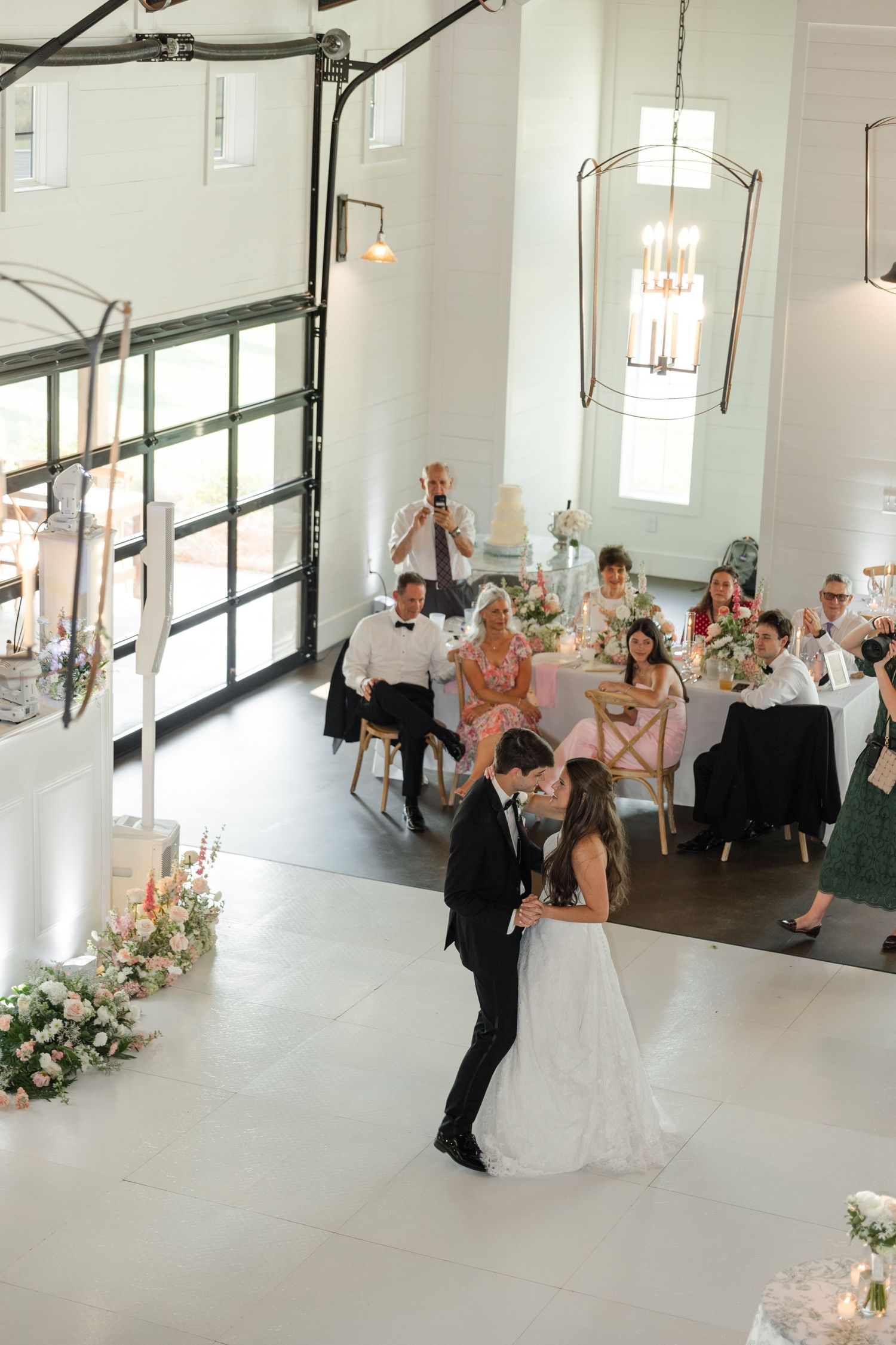 Aerial view of bride and groom sharing first dance in bright white barn wedding venue.