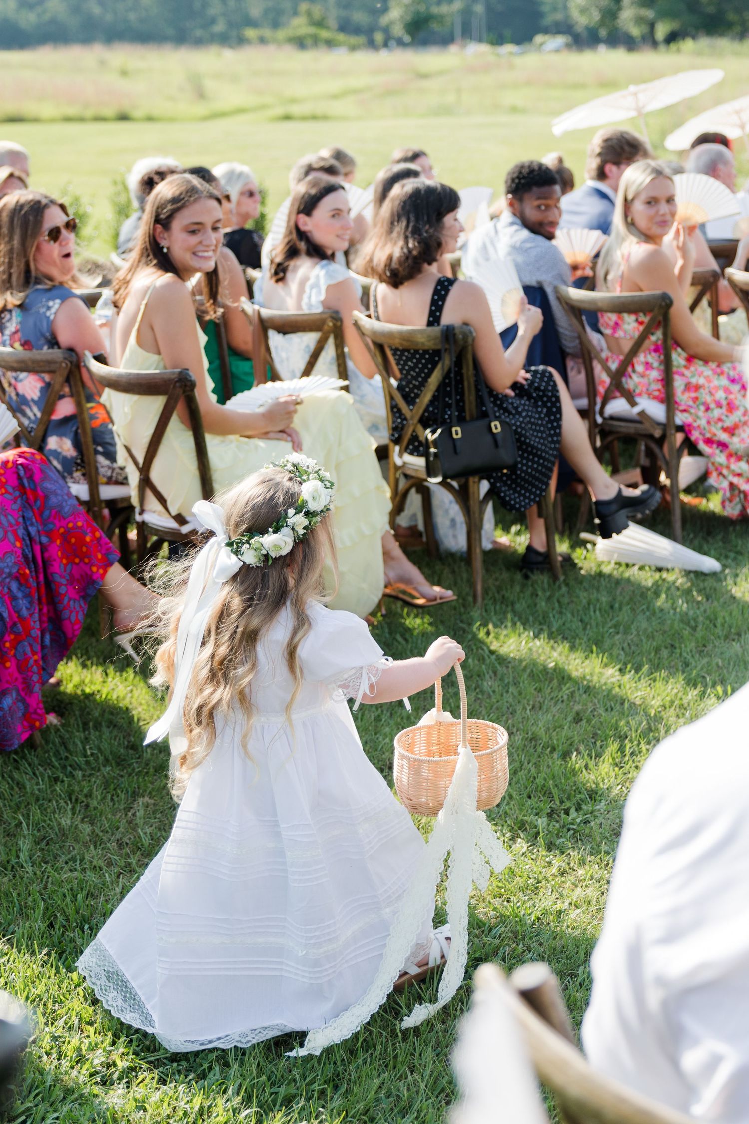 Flower girl in white dress with floral crown carries basket down outdoor wedding ceremony aisle.