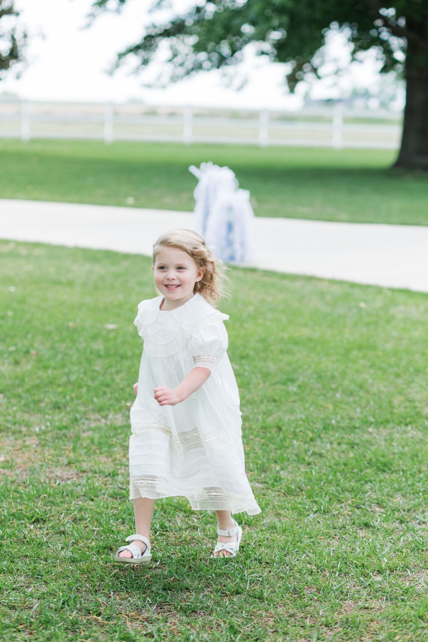 Young flower girl in white dress running on green grass at outdoor wedding ceremony.