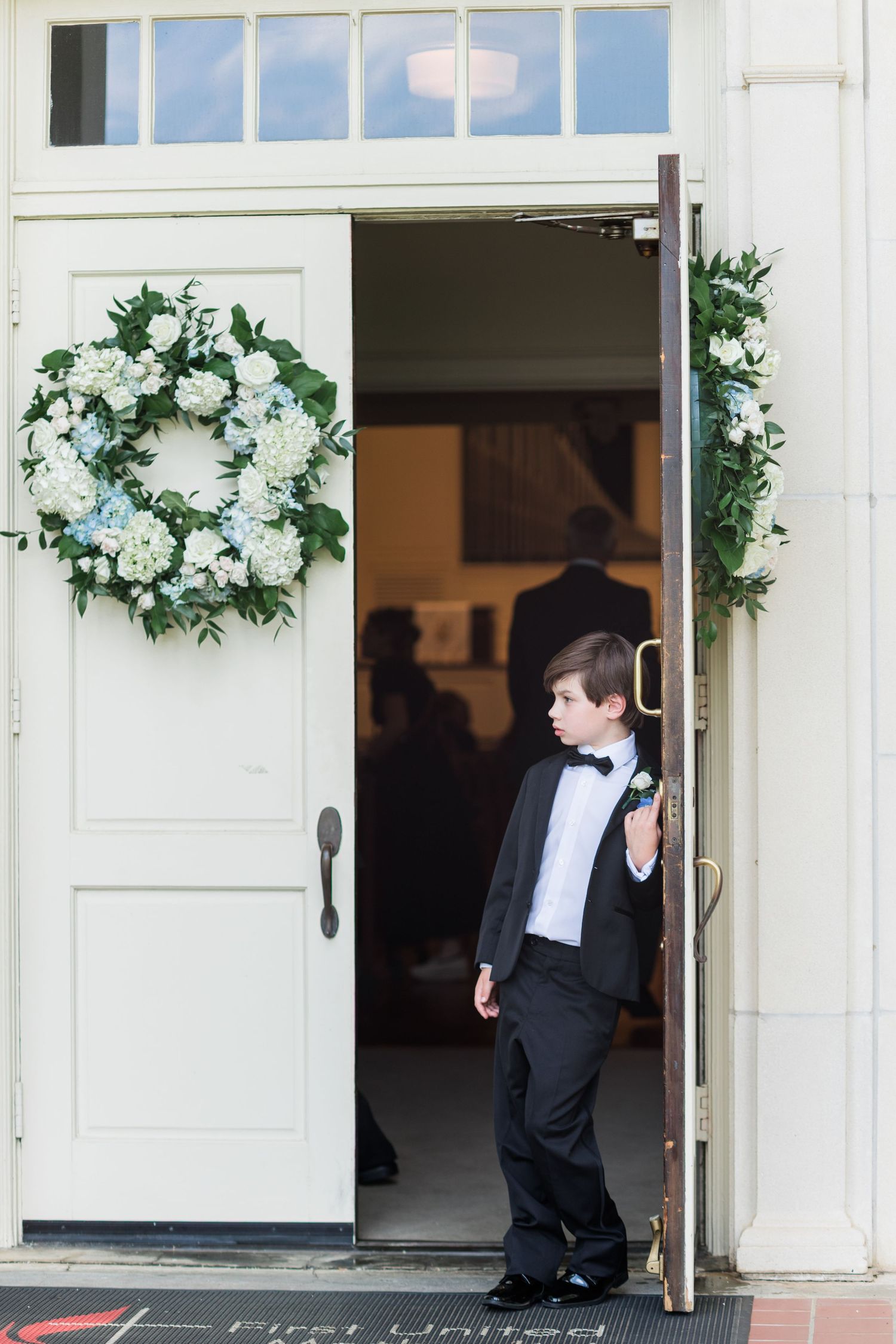 Young ring bearer in black tuxedo standing at church entrance door decorated with white and blue floral wreaths.