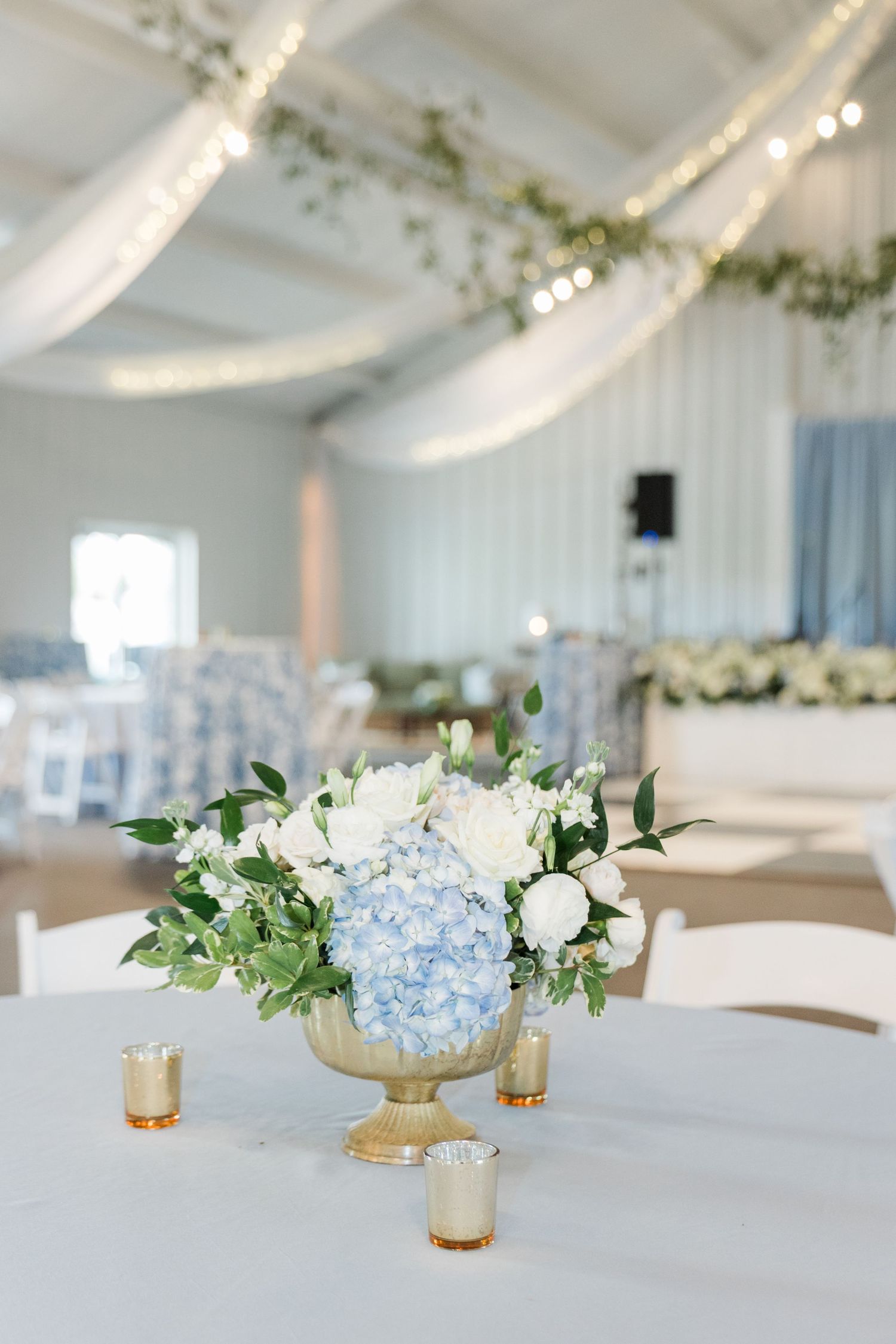 Blue hydrangea and white rose centerpiece in gold urn on white tablecloth at wedding reception.