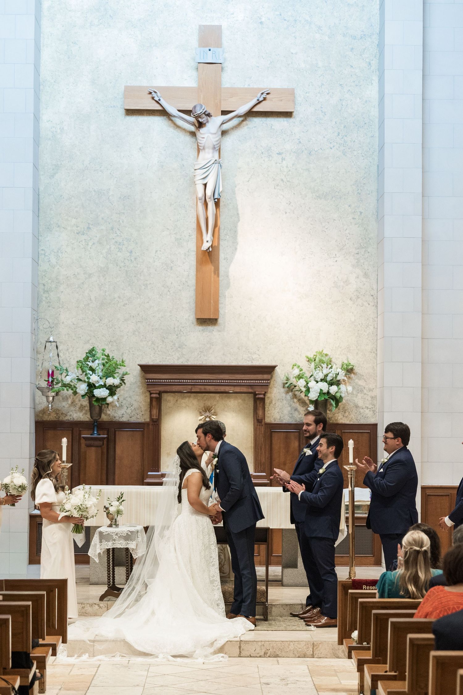Bride and groom share first kiss at Catholic church altar with crucifix, flanked by wedding party during ceremony.