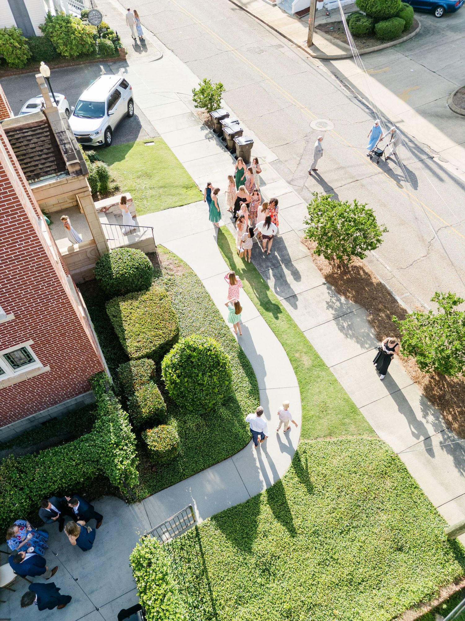 Aerial view of a wedding party walking along a path beside a brick building with manicured hedges.