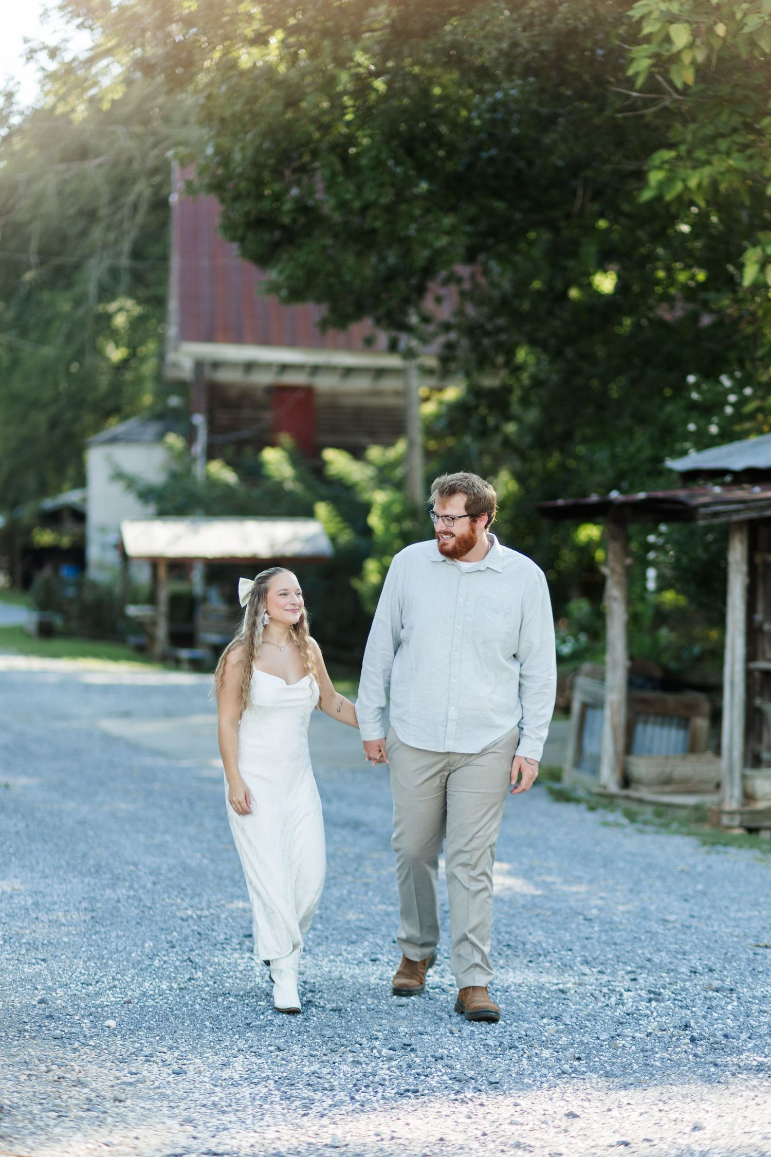 Couple holding hands walking on gravel path past rustic buildings and green trees in warm sunlight.
