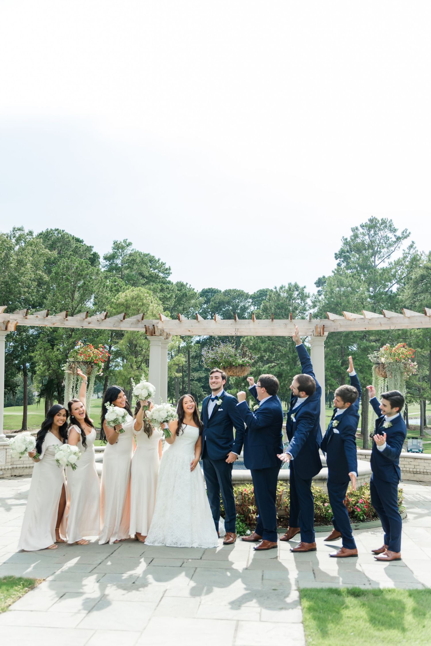 Wedding party celebrating outdoors with bridesmaids in white gowns and groomsmen in navy suits under pergola.