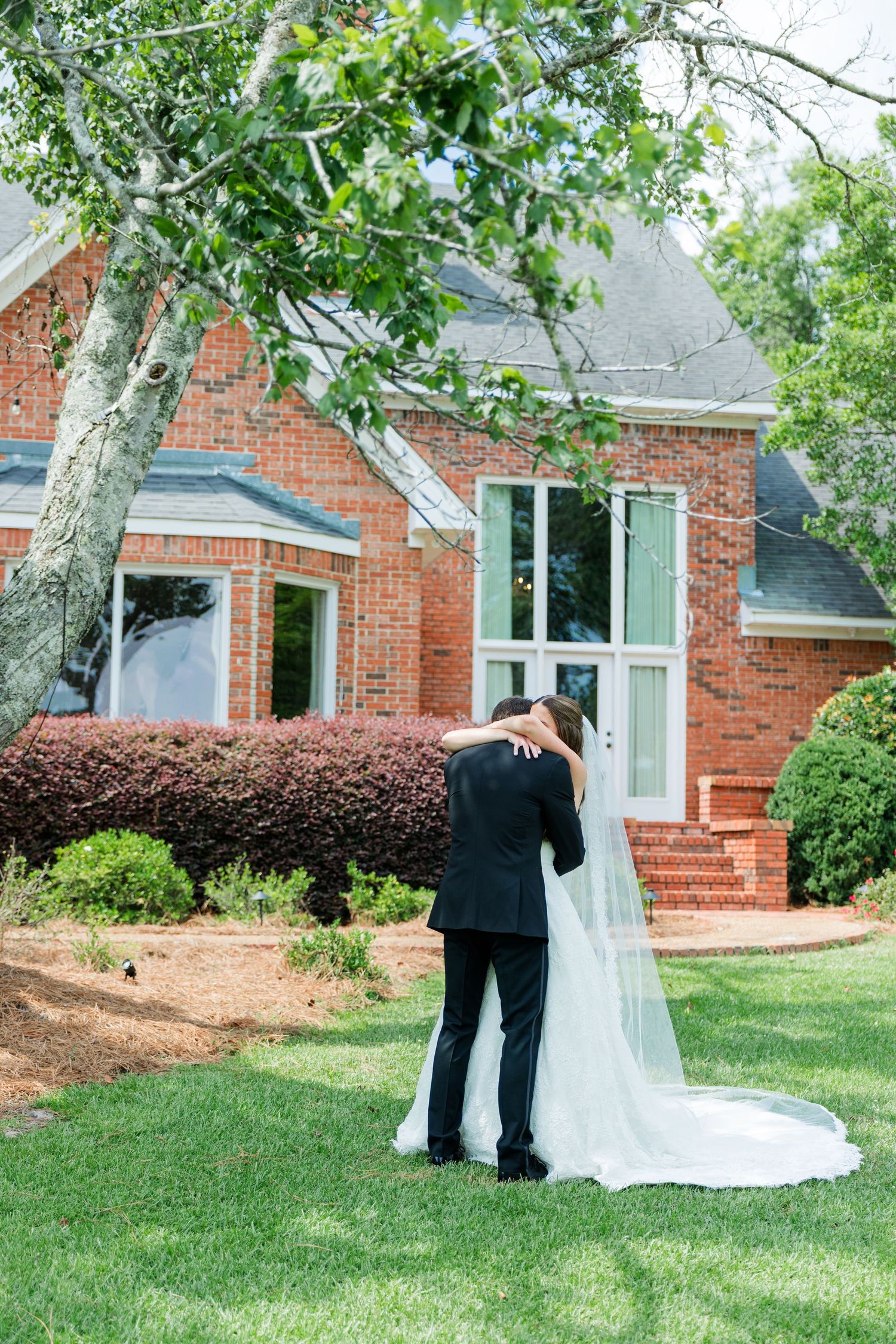 Bride and groom share a tender embrace during first look outside a brick building with lush greenery.