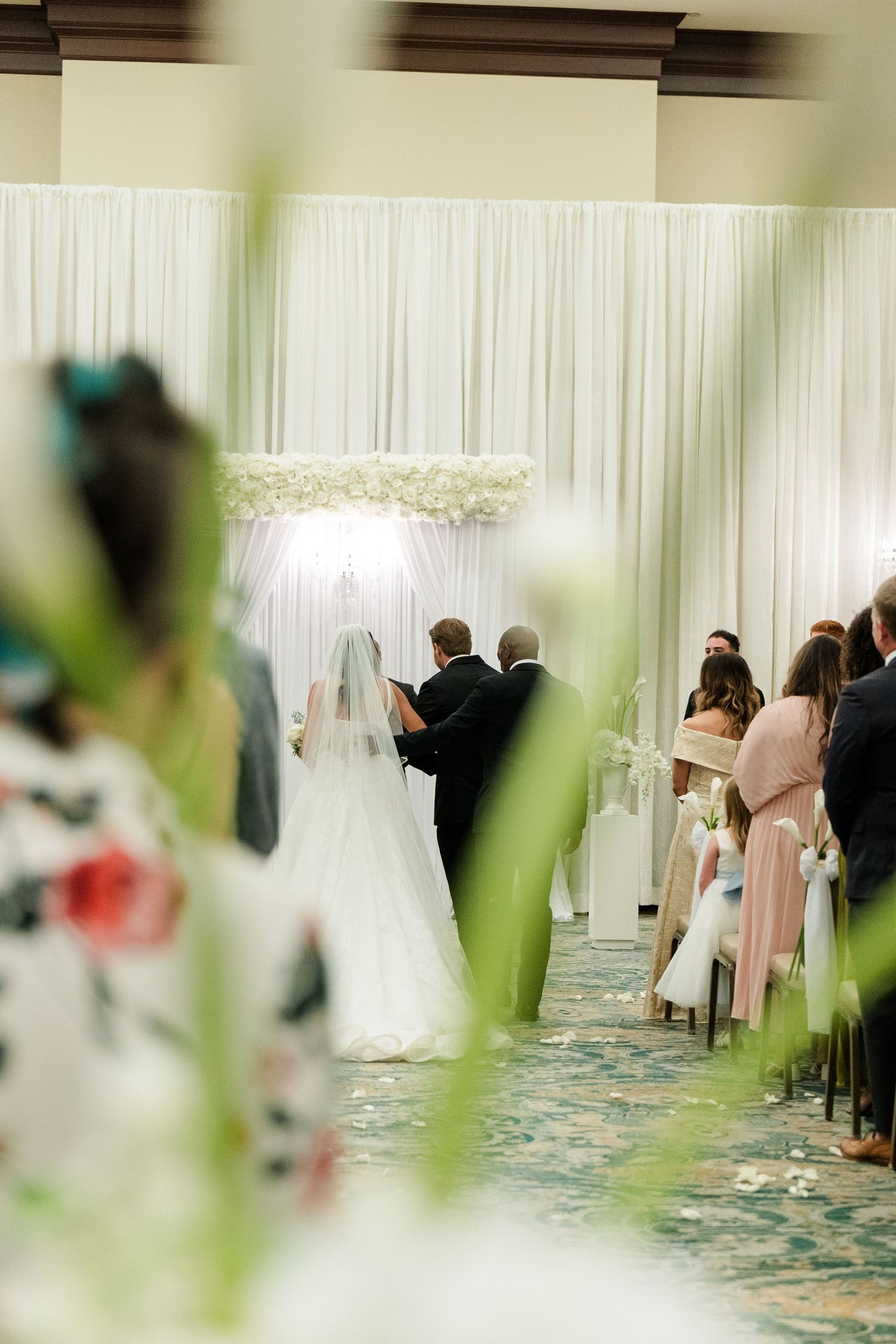 Elegant indoor wedding ceremony with bride and groom at floral arch altar, guests seated in aisle, white draping backdrop.