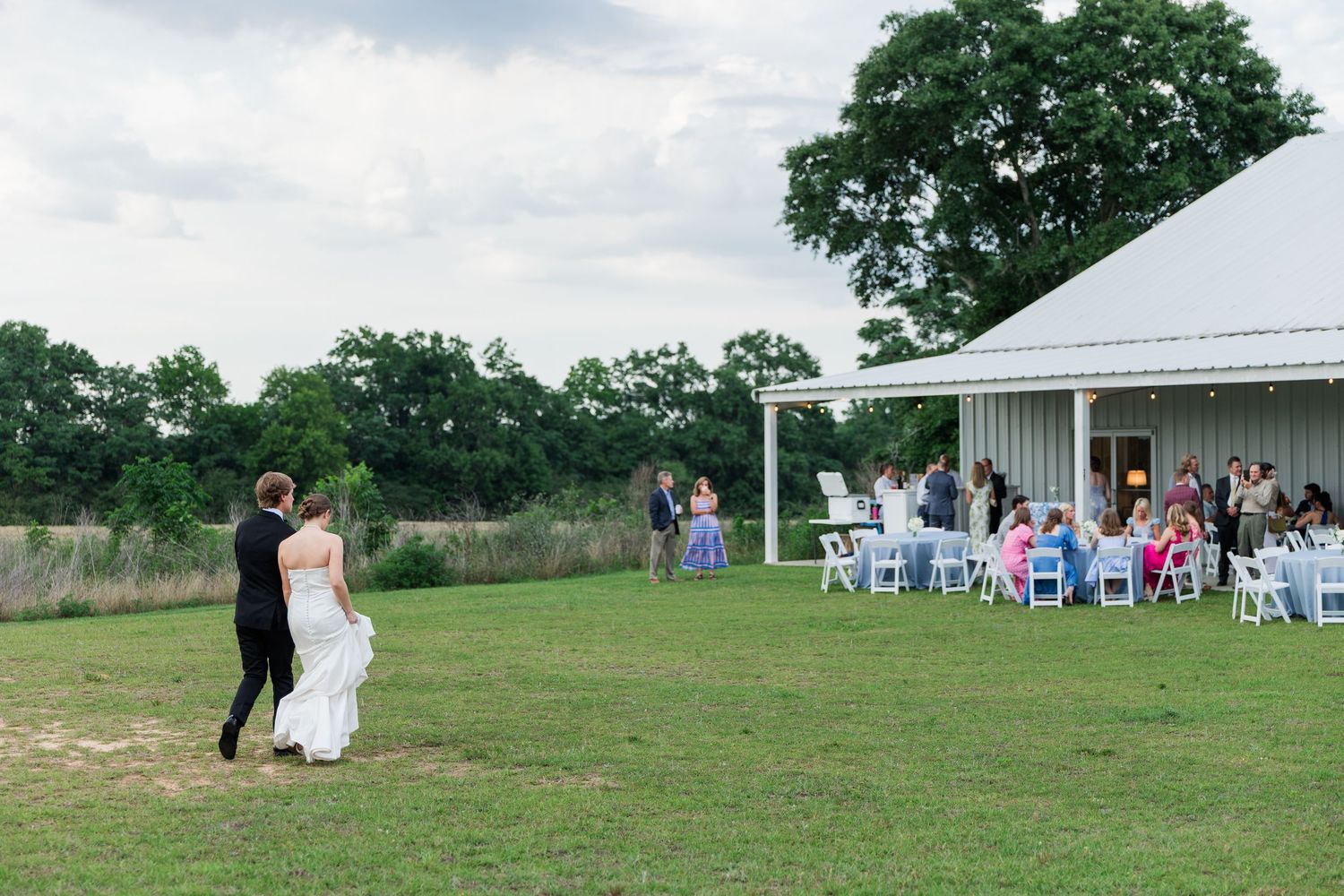 Bride and groom walking toward outdoor wedding reception tent on green lawn at sunset.