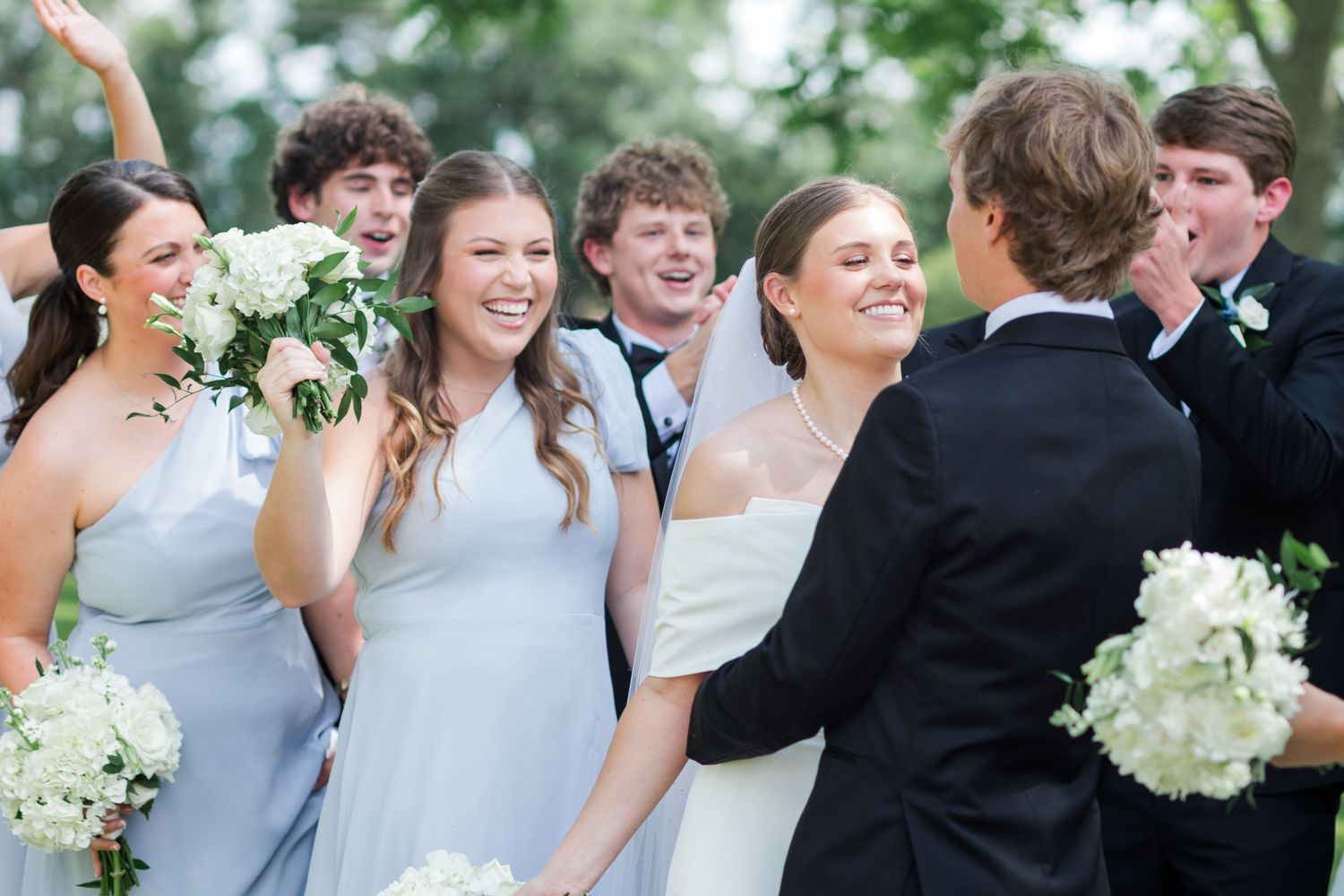 Bride and groom sharing a joyful moment with bridesmaids in blue dresses and groomsmen celebrating at outdoor wedding.