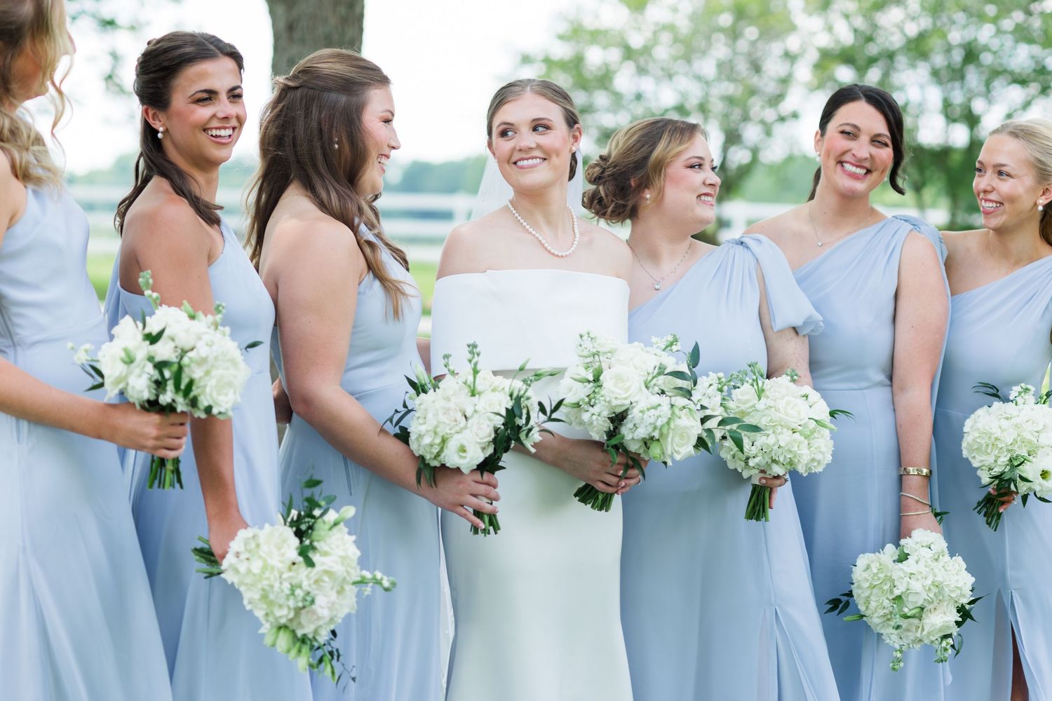 Bride in white gown surrounded by bridesmaids in light blue dresses holding white floral bouquets.