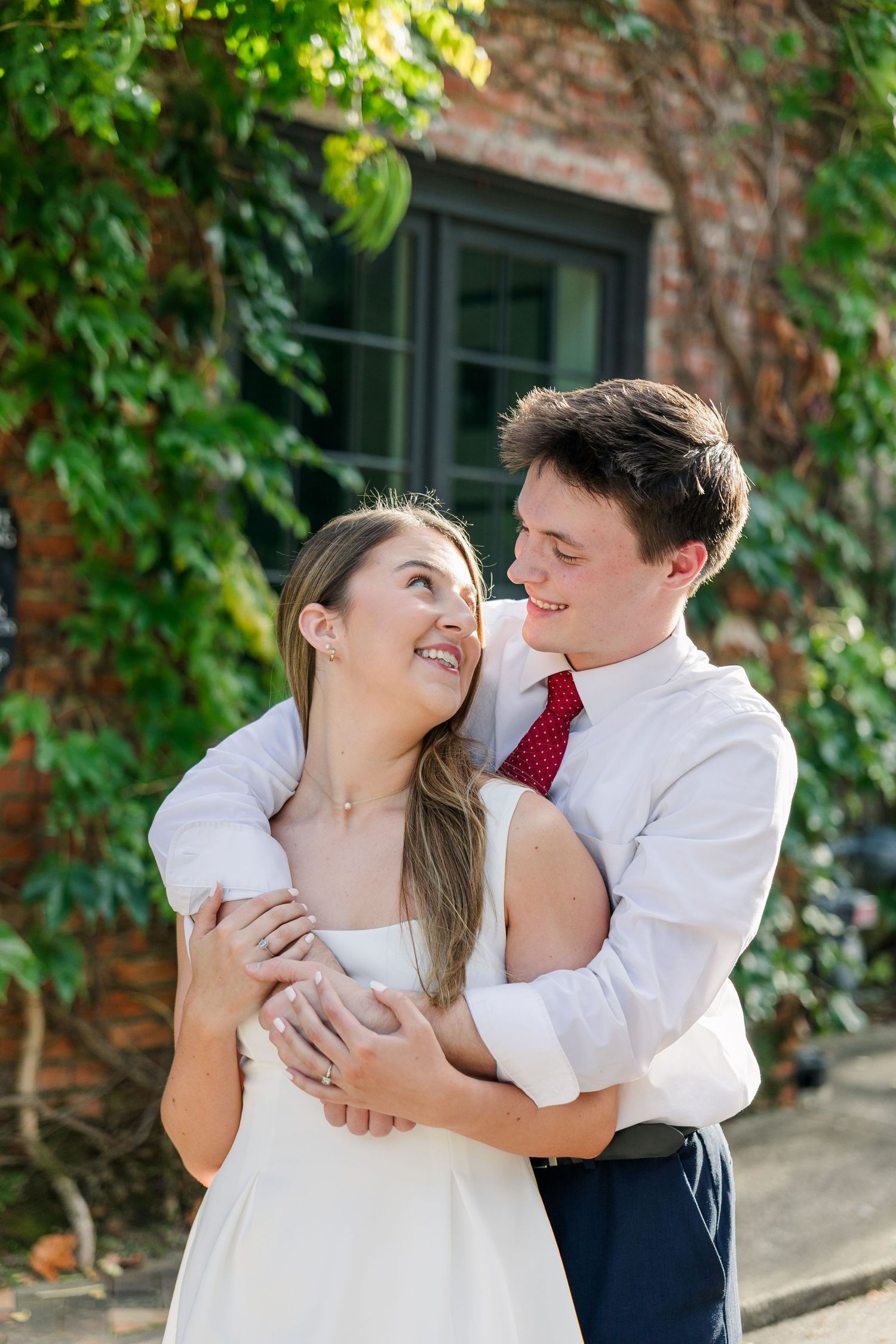 Smiling couple embracing outdoors near ivy-covered brick wall, woman in white dress, man in white shirt and red tie.
