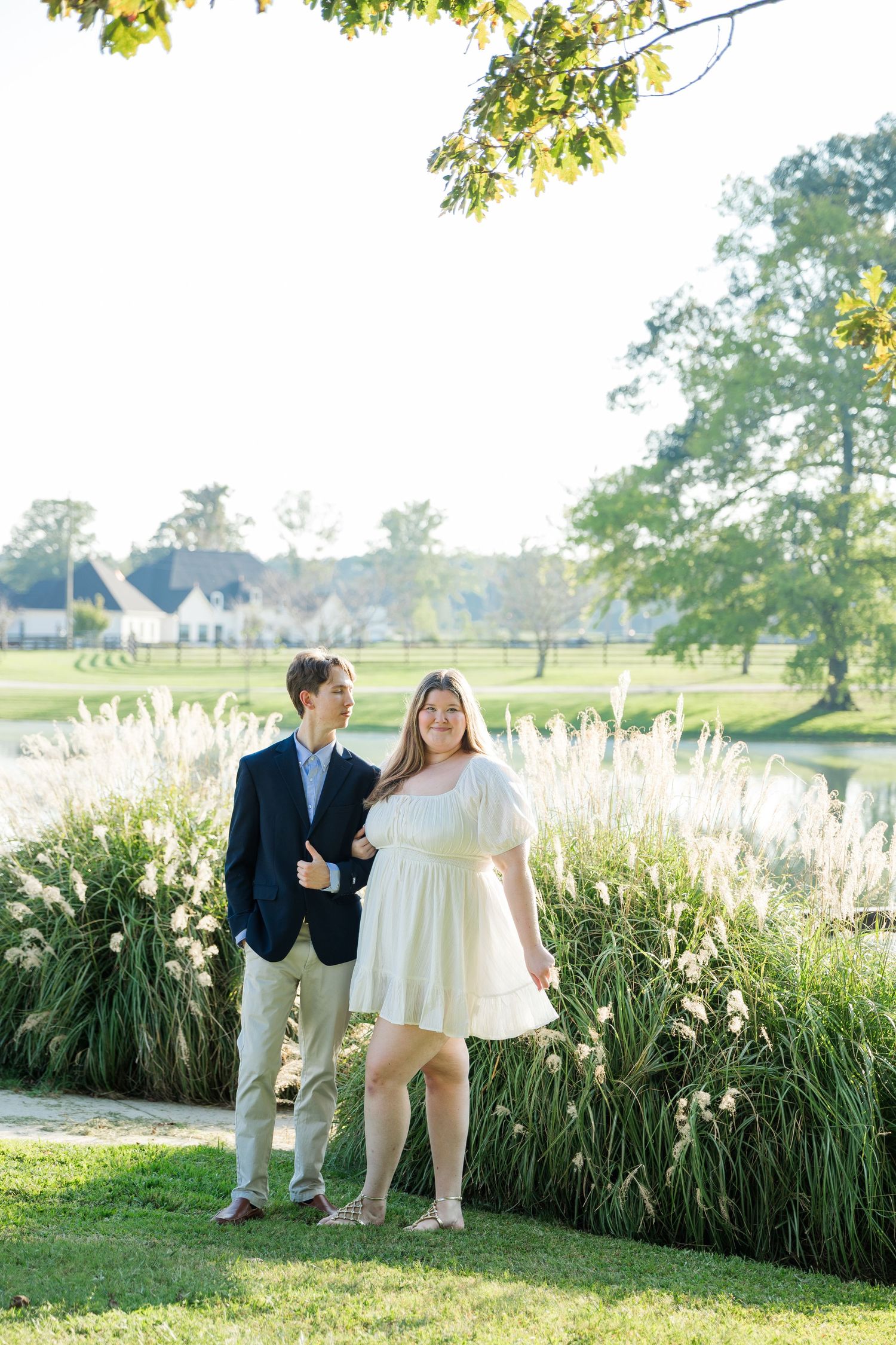 Couple posing together near tall ornamental grasses by a pond, man in navy blazer and woman in white mini dress.