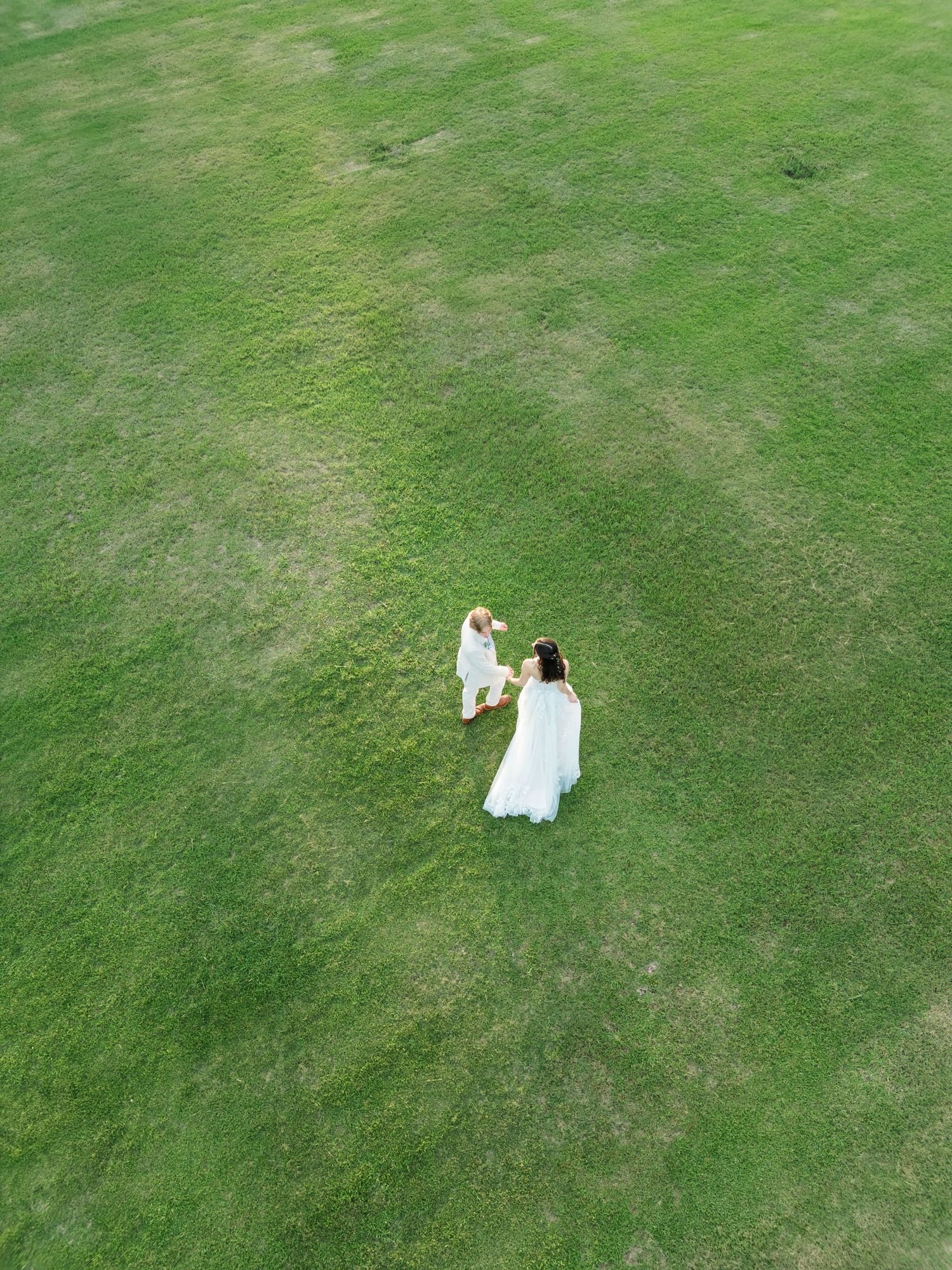 Aerial drone view of a woman in a white dress holding hands with a child on a lush green grass field.