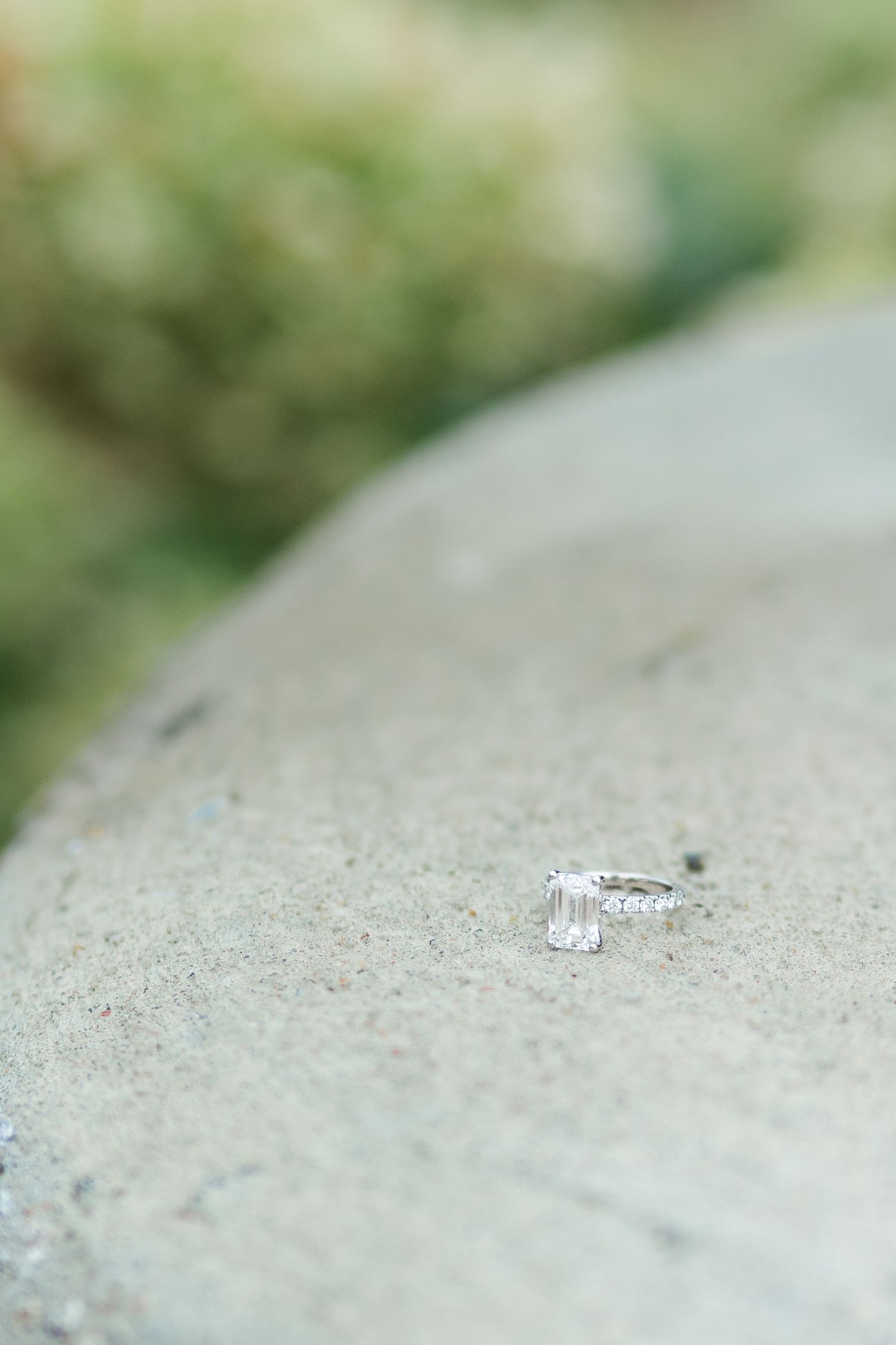 Elegant emerald cut diamond engagement ring and wedding band resting on a smooth stone surface outdoors.