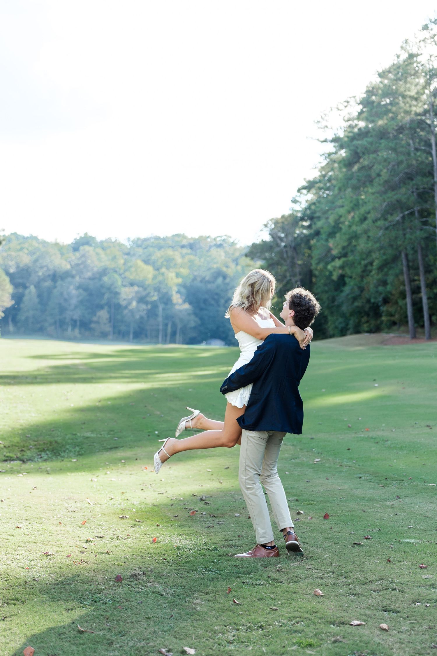 Groom lifting bride on a sunny golf course fairway, both laughing joyfully surrounded by lush trees.