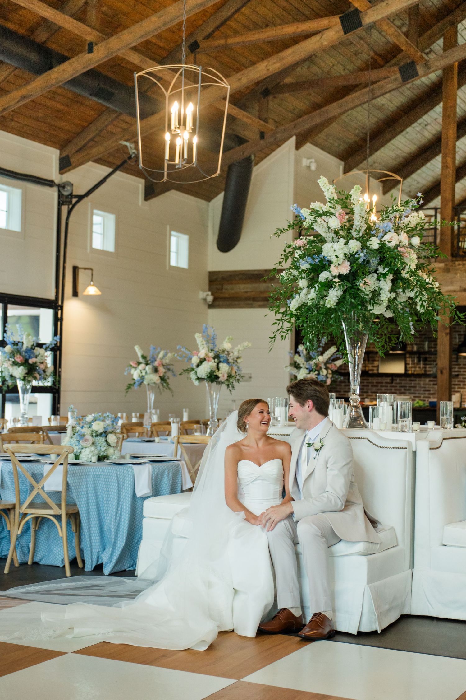 Bride and groom sit together at elegant wedding reception with blue linens and tall floral arrangements in rustic barn venue.