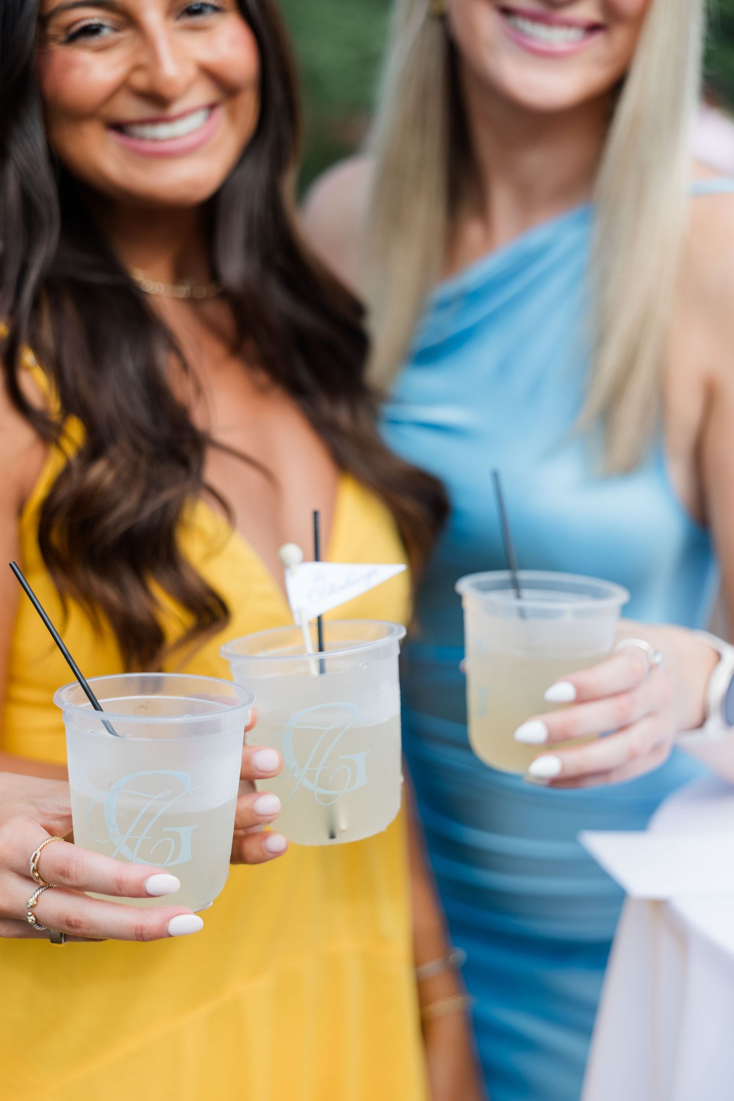Two smiling women in yellow and blue dresses toasting with lemonade cocktails in plastic cups at an outdoor event.
