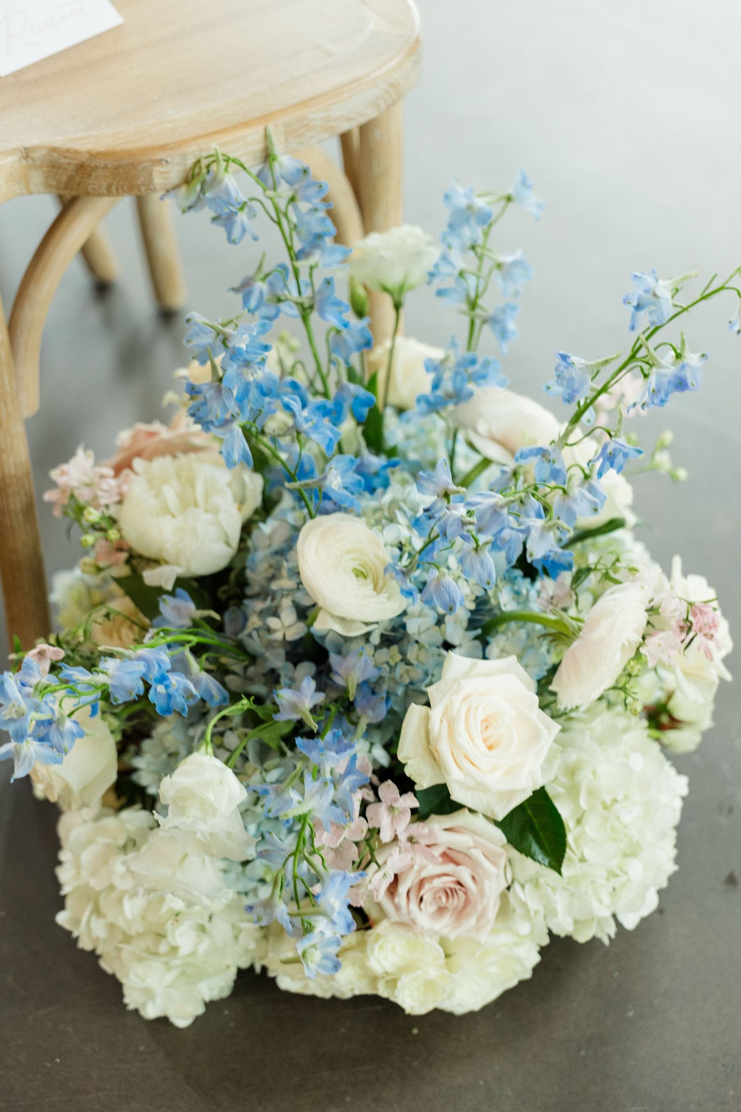 Elegant floral arrangement with white hydrangeas, cream roses, blush pink roses, and blue delphinium on a gray floor.