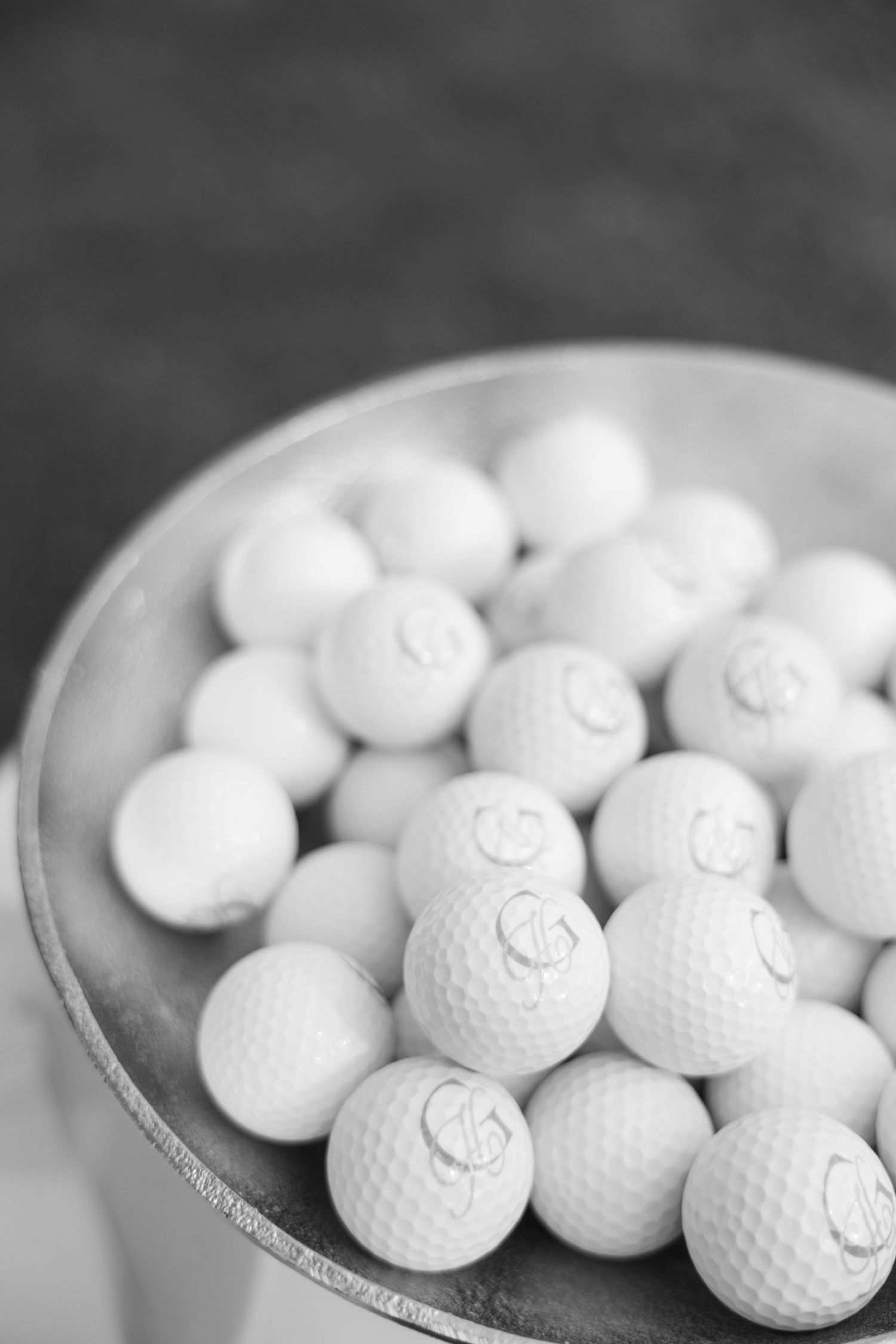 Black and white photo of golf balls piled in a metal bowl, showing dimpled texture and monogram markings.