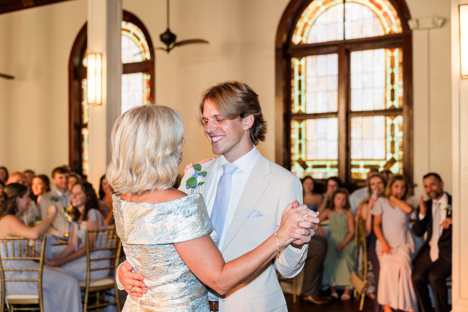 Groom in a tan suit sharing a mother-son dance in an elegant wedding reception hall.