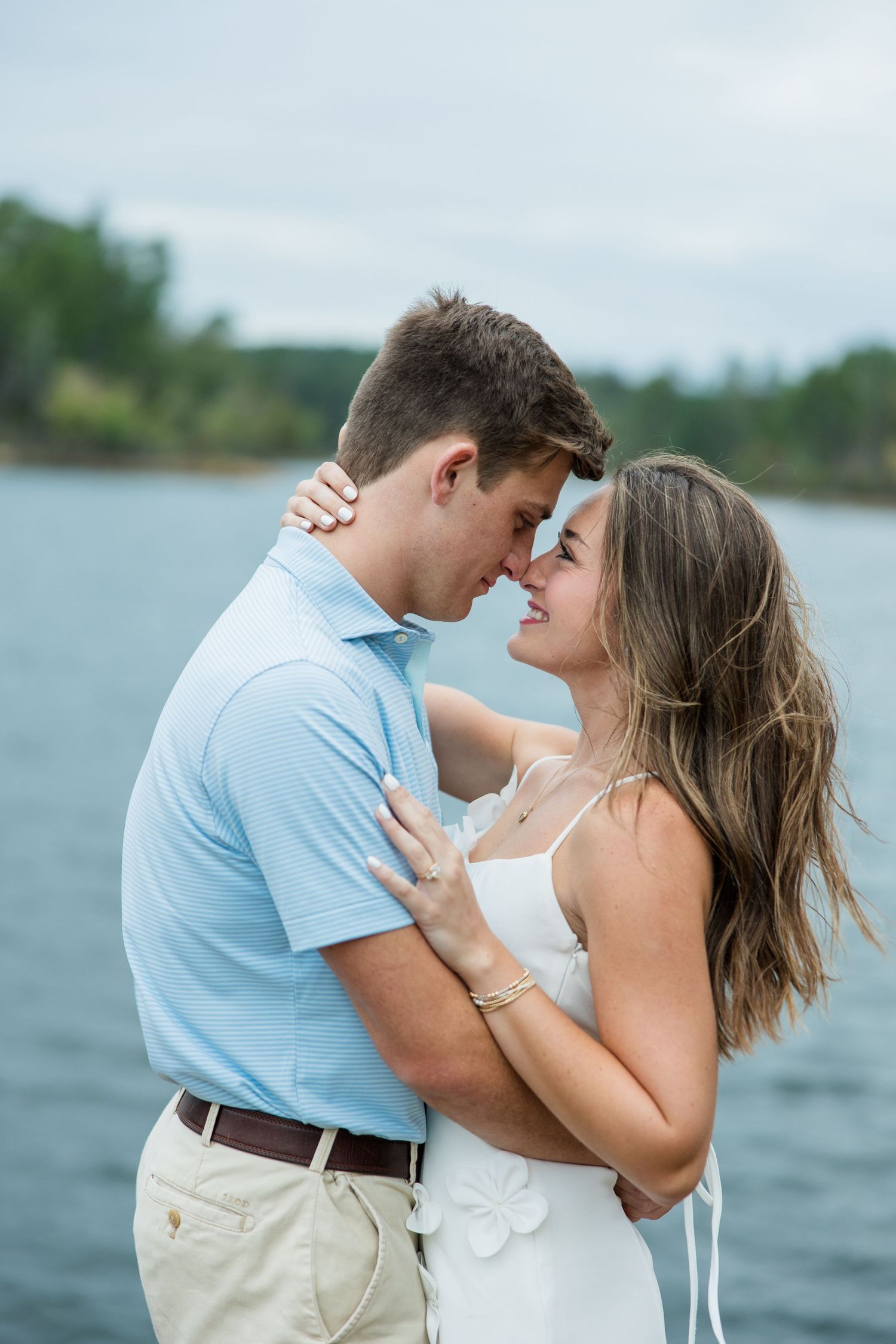 Smiling couple in an intimate embrace, touching noses by a calm lake with tree-lined shores, in an engagement photo.
