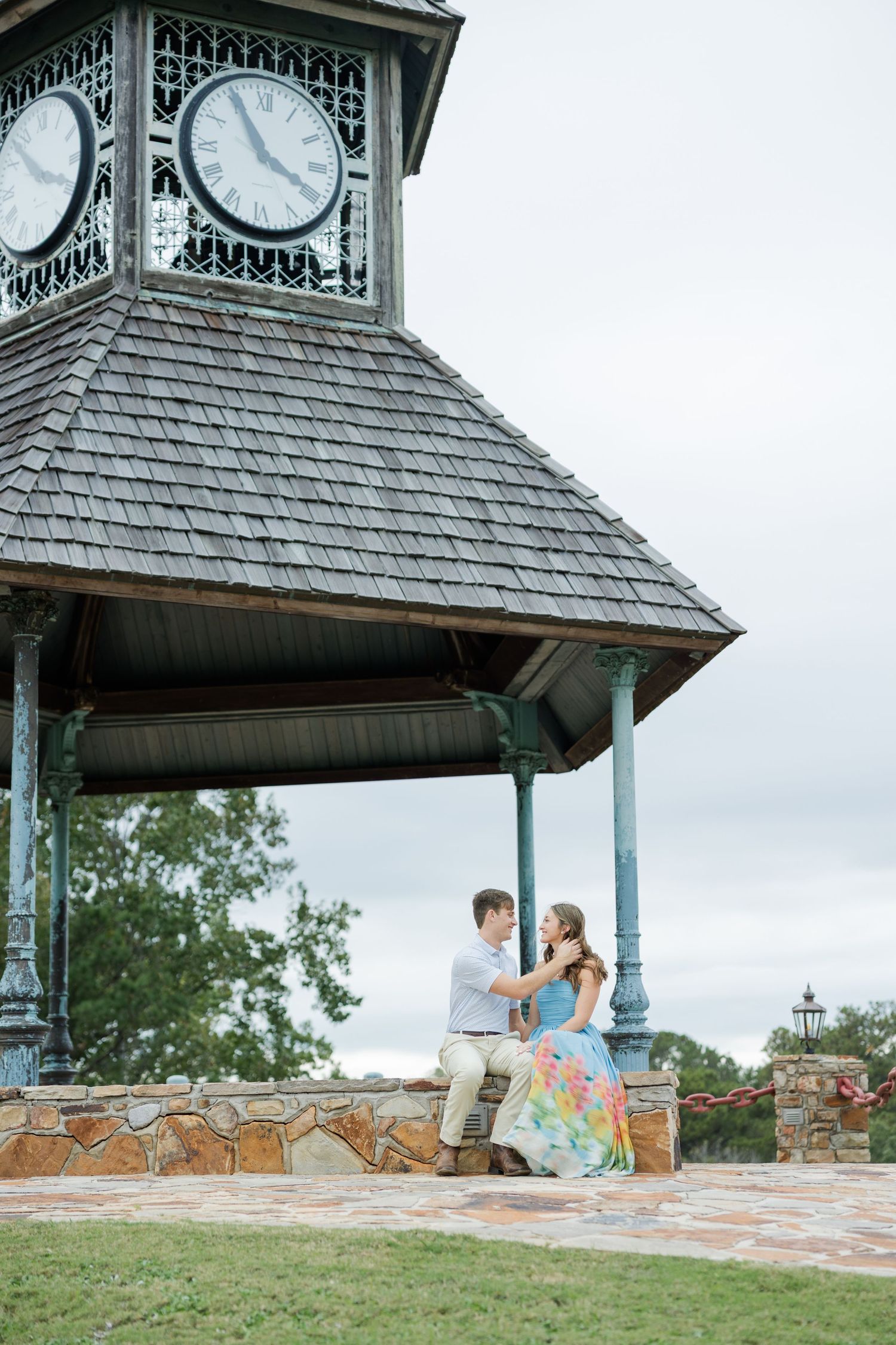 Engaged couple sitting close together under a clock tower pavilion with a stone wall during an outdoor engagement photo session.