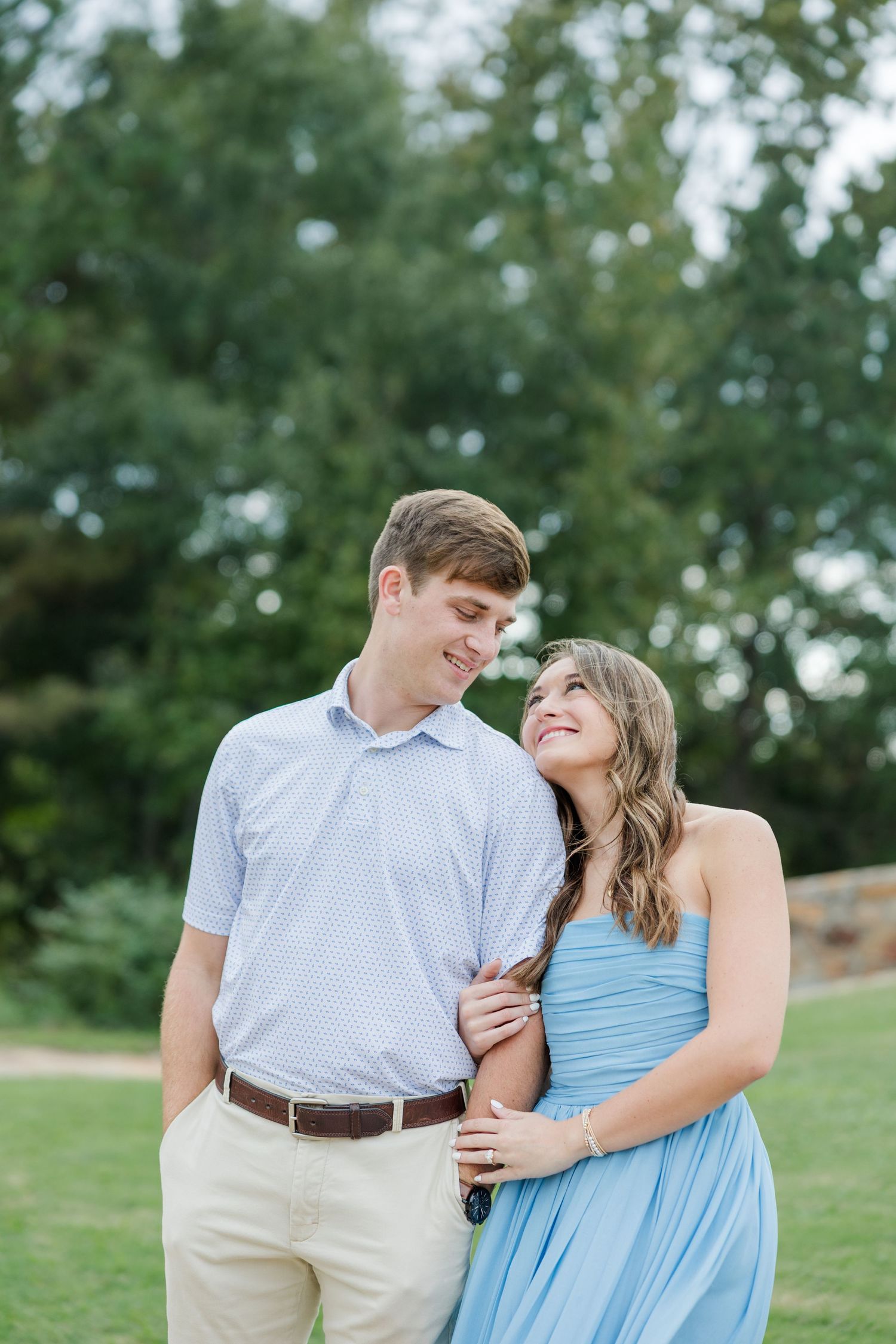 Smiling couple in coordinated outfits embrace outdoors, the woman in a light blue dress and the man in a polo, with green trees behind.
