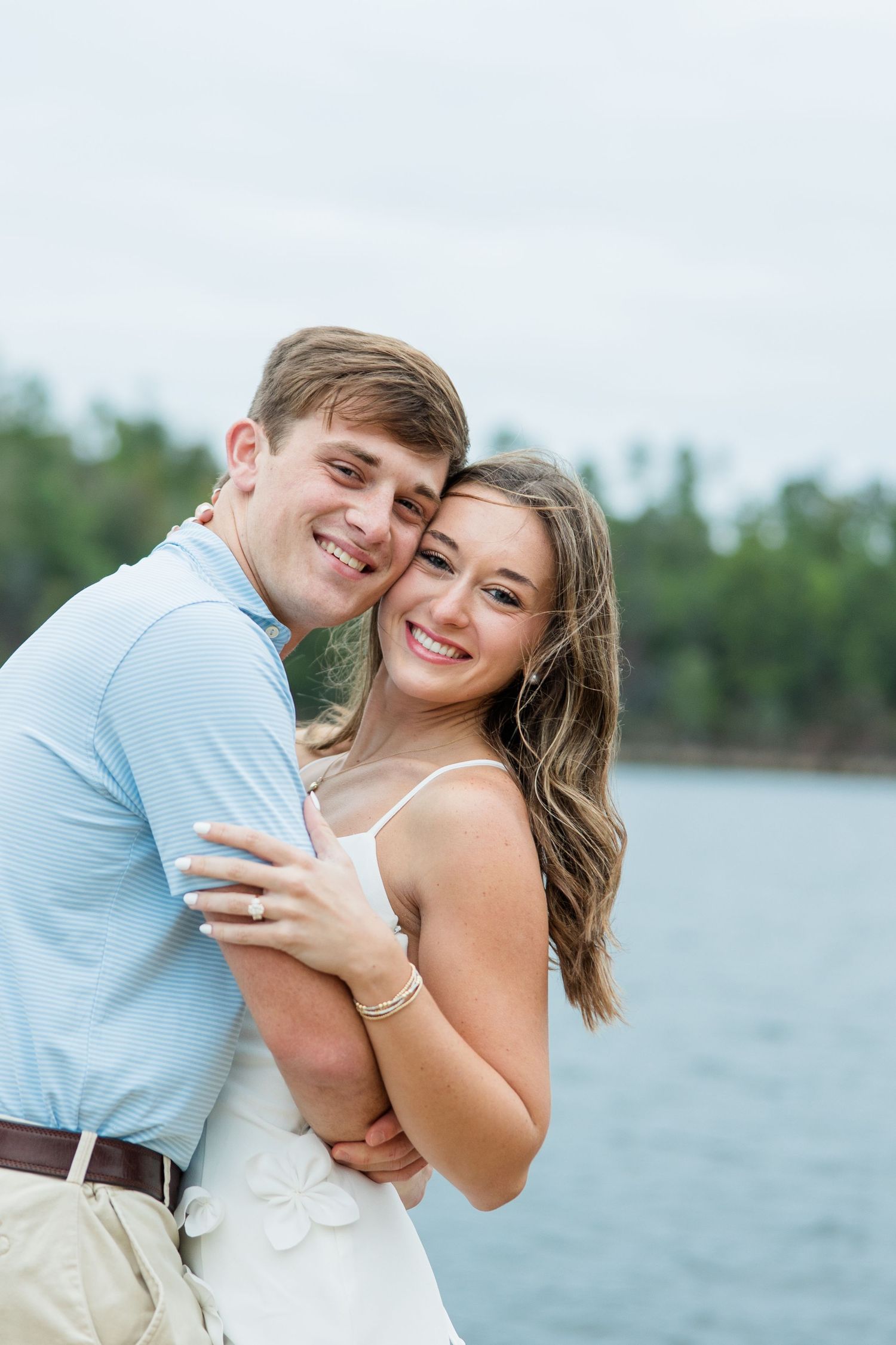 Smiling engaged couple hugging and posing cheek to cheek beside a lake with a tree-lined shore and a cloudy sky.