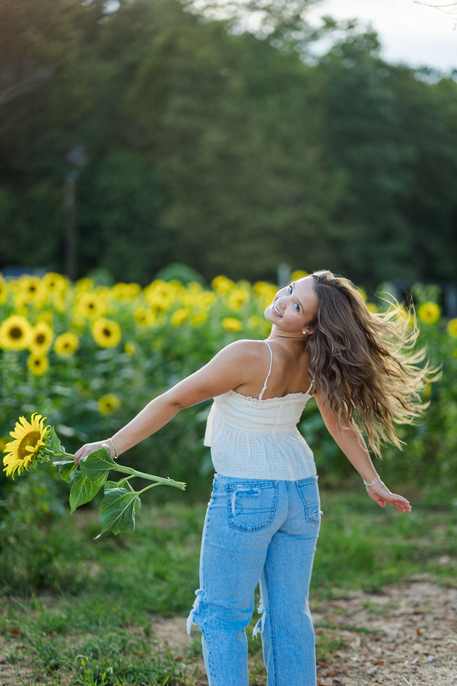 A joyful woman twirls in a sunflower field, holding a sunflower, wearing a white top and jeans, with hair flowing.