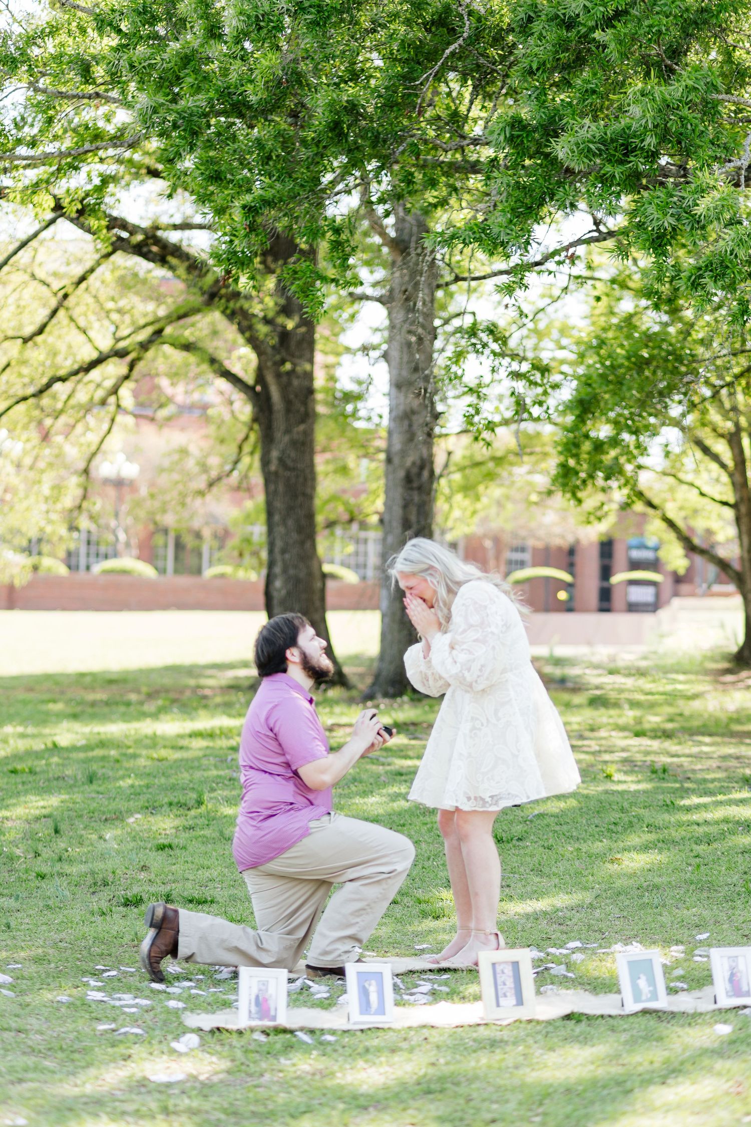 Man kneeling on one knee proposing to emotional woman in white dress outdoors in a park surrounded by photos.