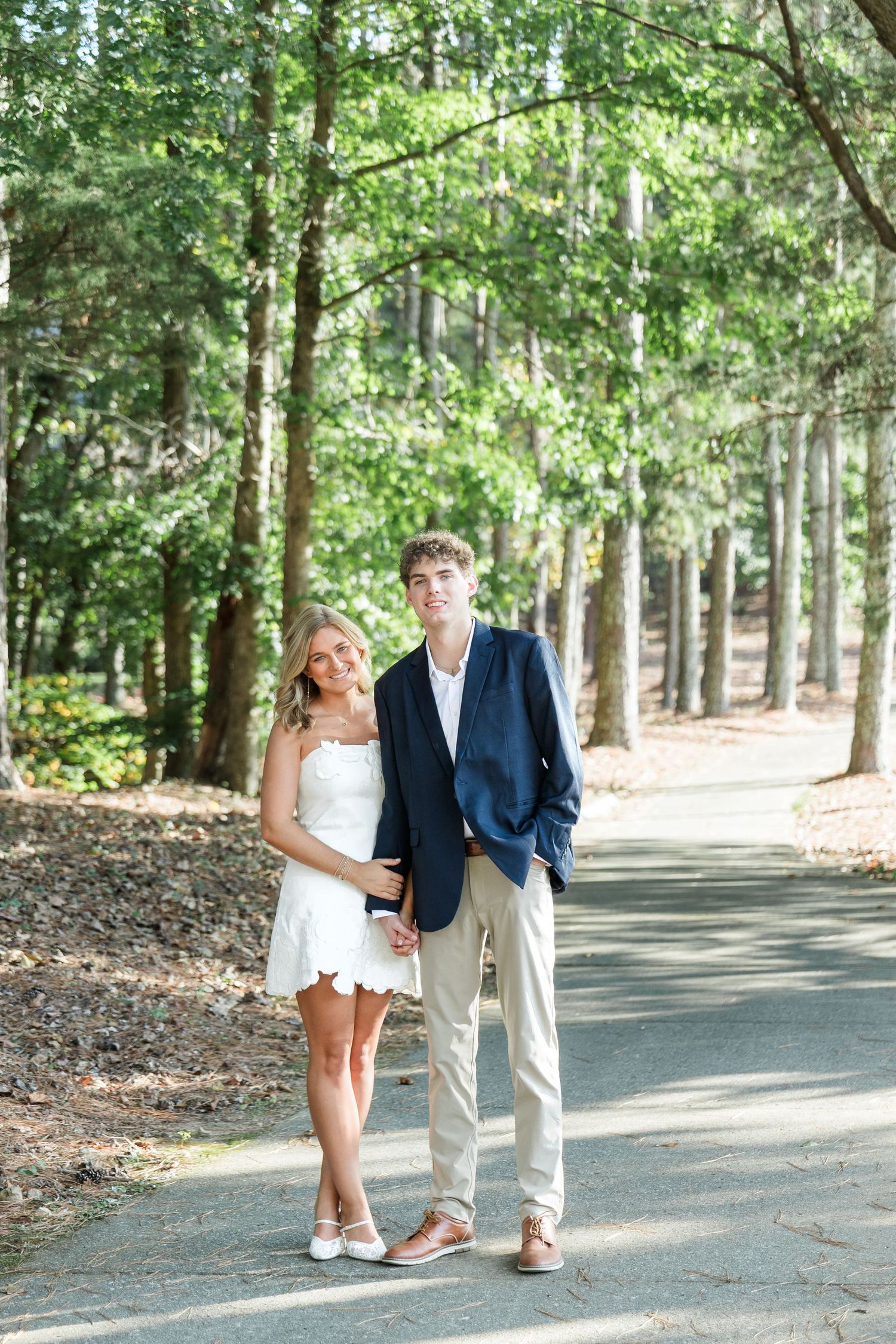 Engaged couple holding hands on a tree-lined path, bride in white dress and groom in navy blazer and khakis.