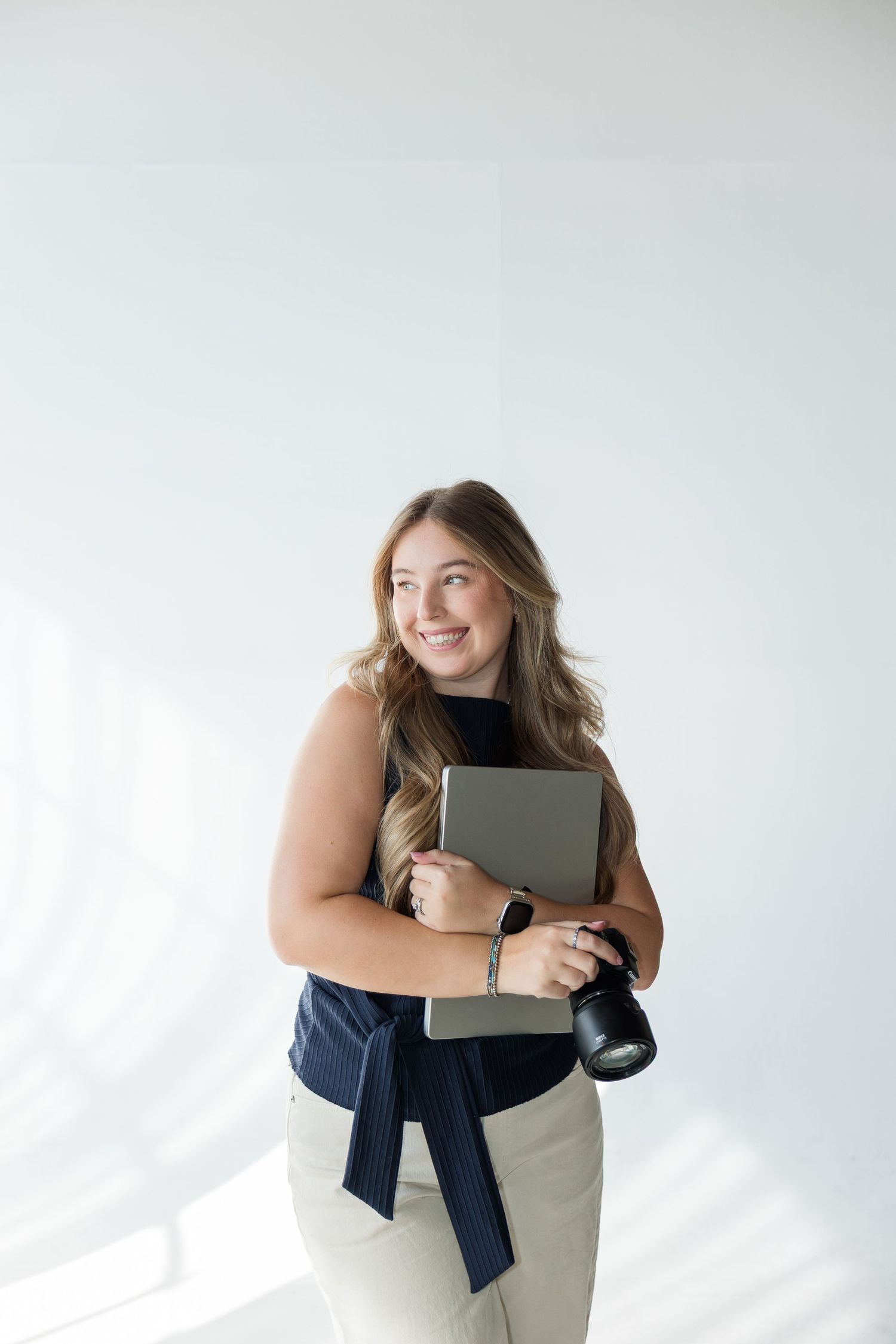 Smiling woman holding a laptop and camera, wearing a navy top, posing against a white background.