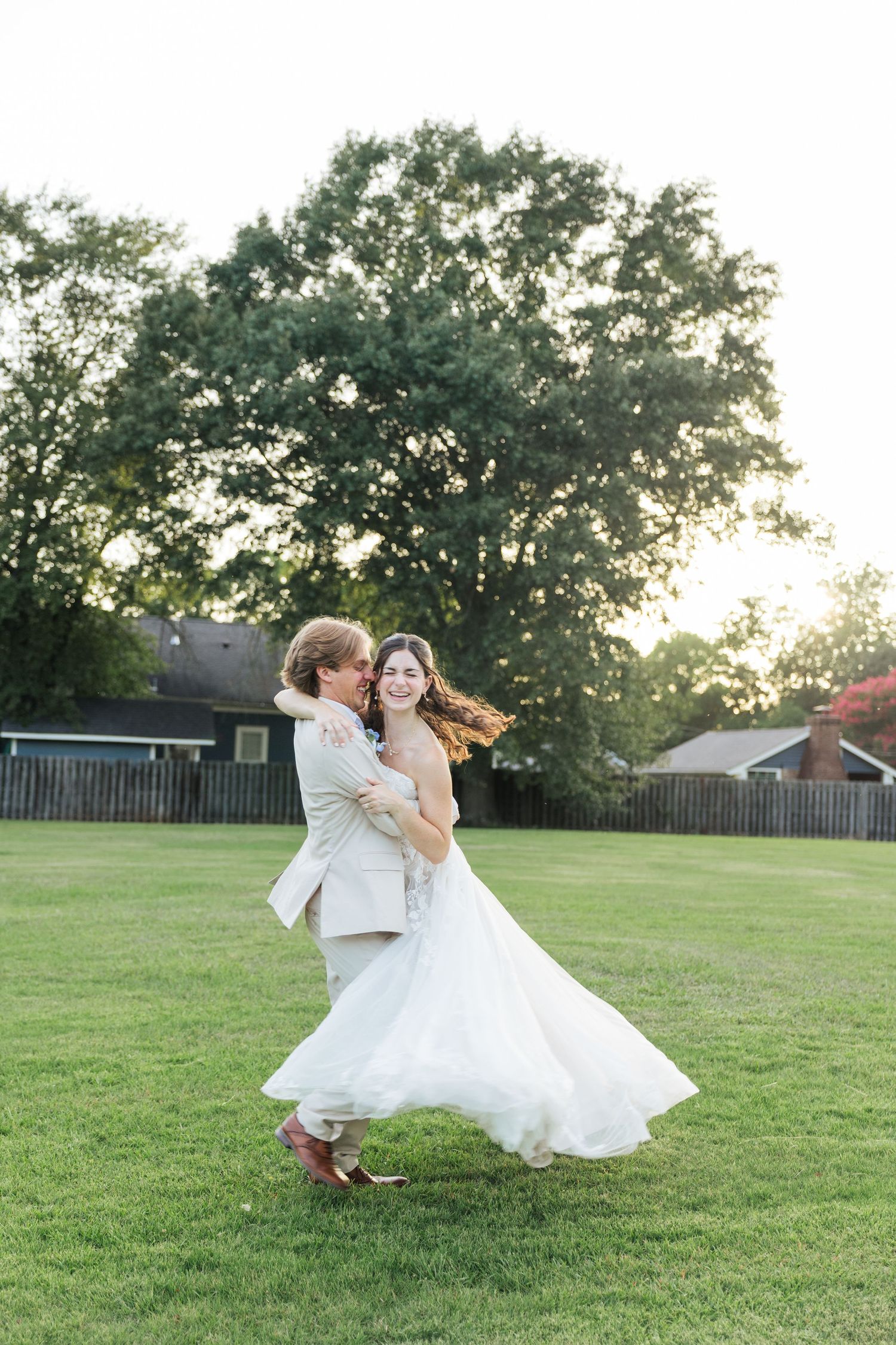 Groom joyfully lifts and spins his bride in a white gown on a green lawn at sunset.