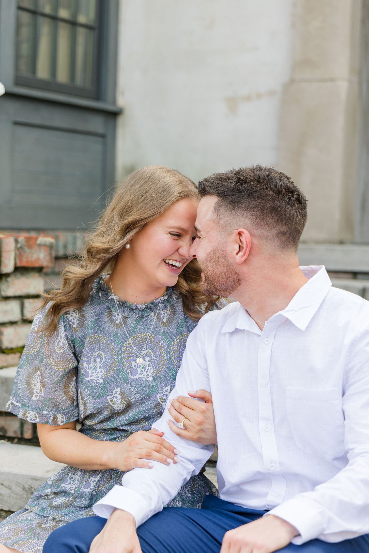 A laughing couple sits close together on stone steps, touching foreheads in a tender moment during an engagement photo session.