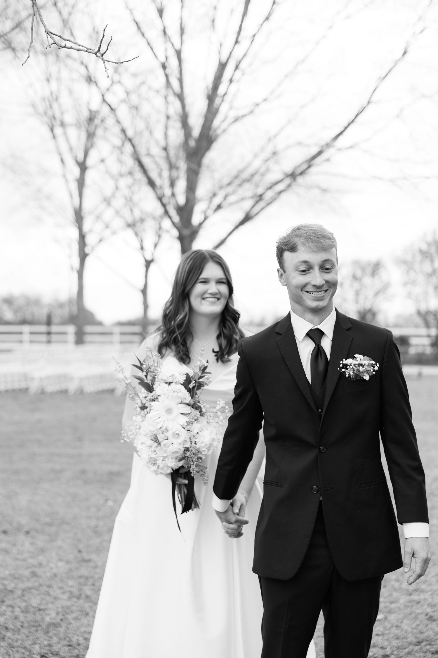 Black and white photo of a smiling bride and groom holding hands outdoors on their wedding day.