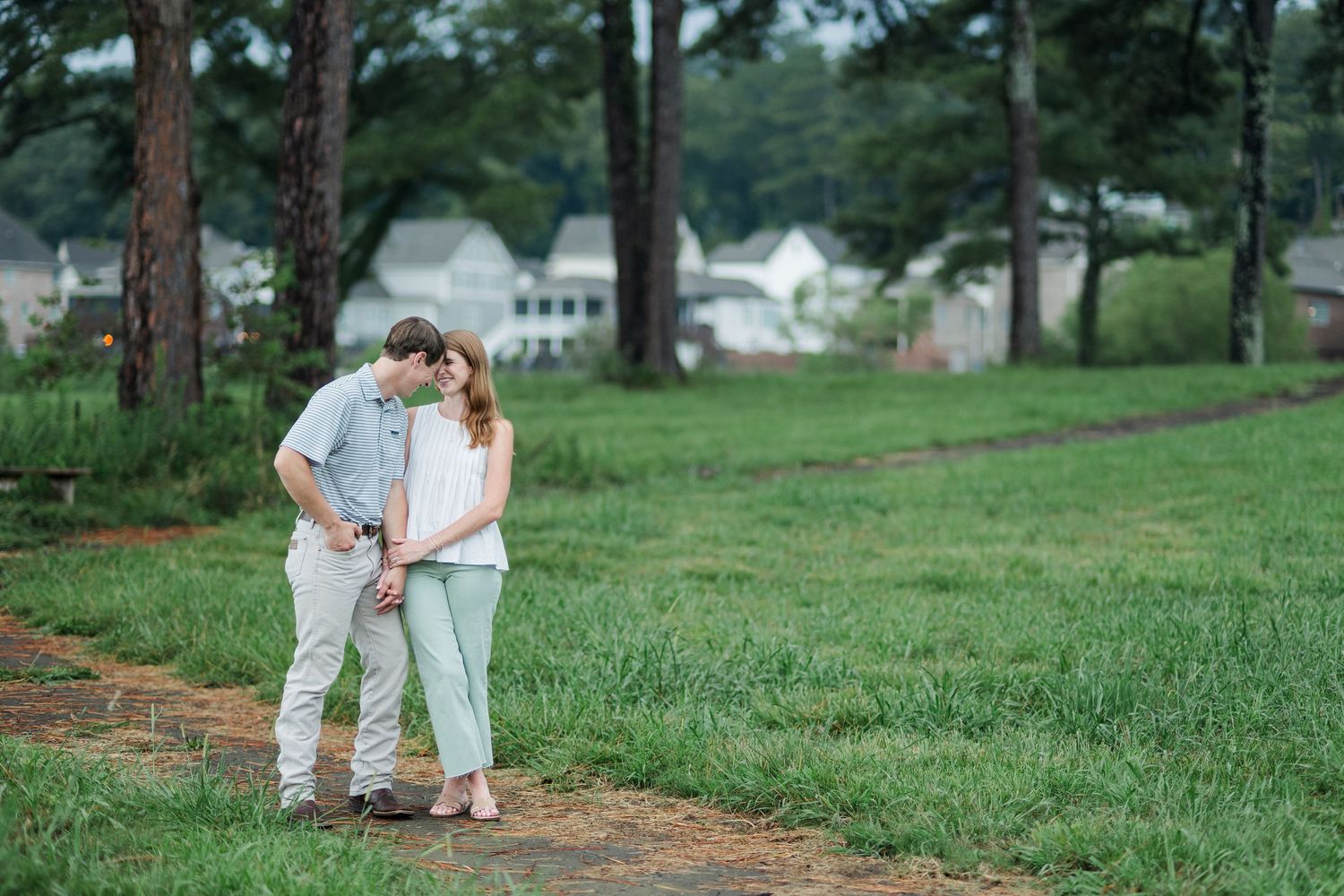 Romantic couple embracing on a garden path surrounded by lush green grass and tall pine trees near suburban homes.