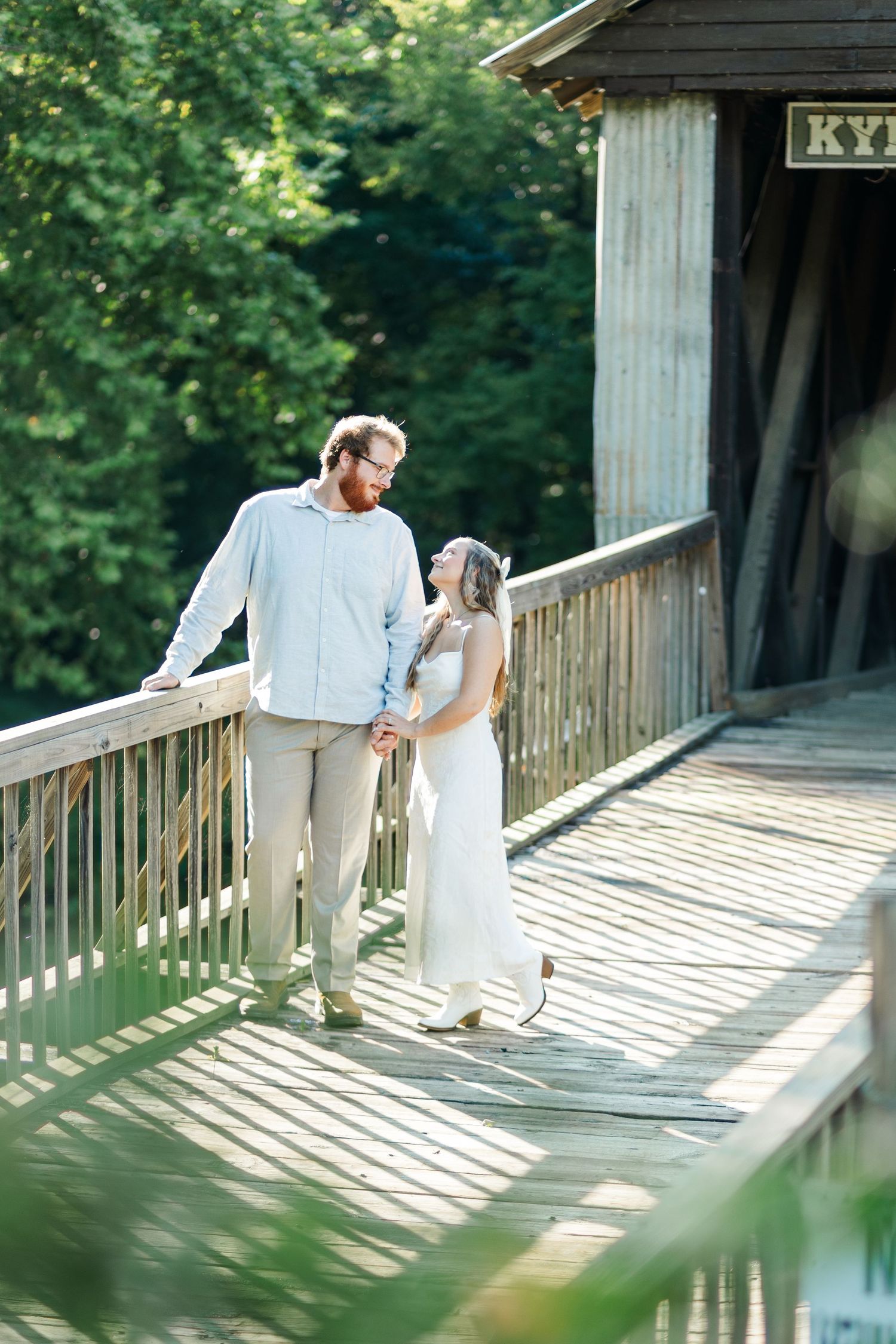 Couple holding hands and gazing at each other on wooden covered bridge with green trees in background.