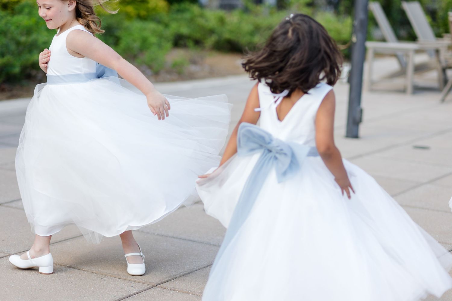 Two flower girls in white tulle dresses with blue sashes twirl and run together at an outdoor wedding venue.