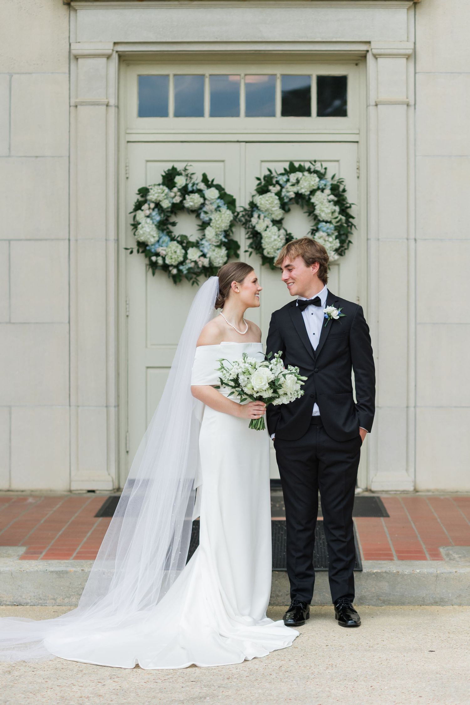 Bride and groom stand before elegant double doors adorned with white floral wreaths at their classic wedding ceremony.