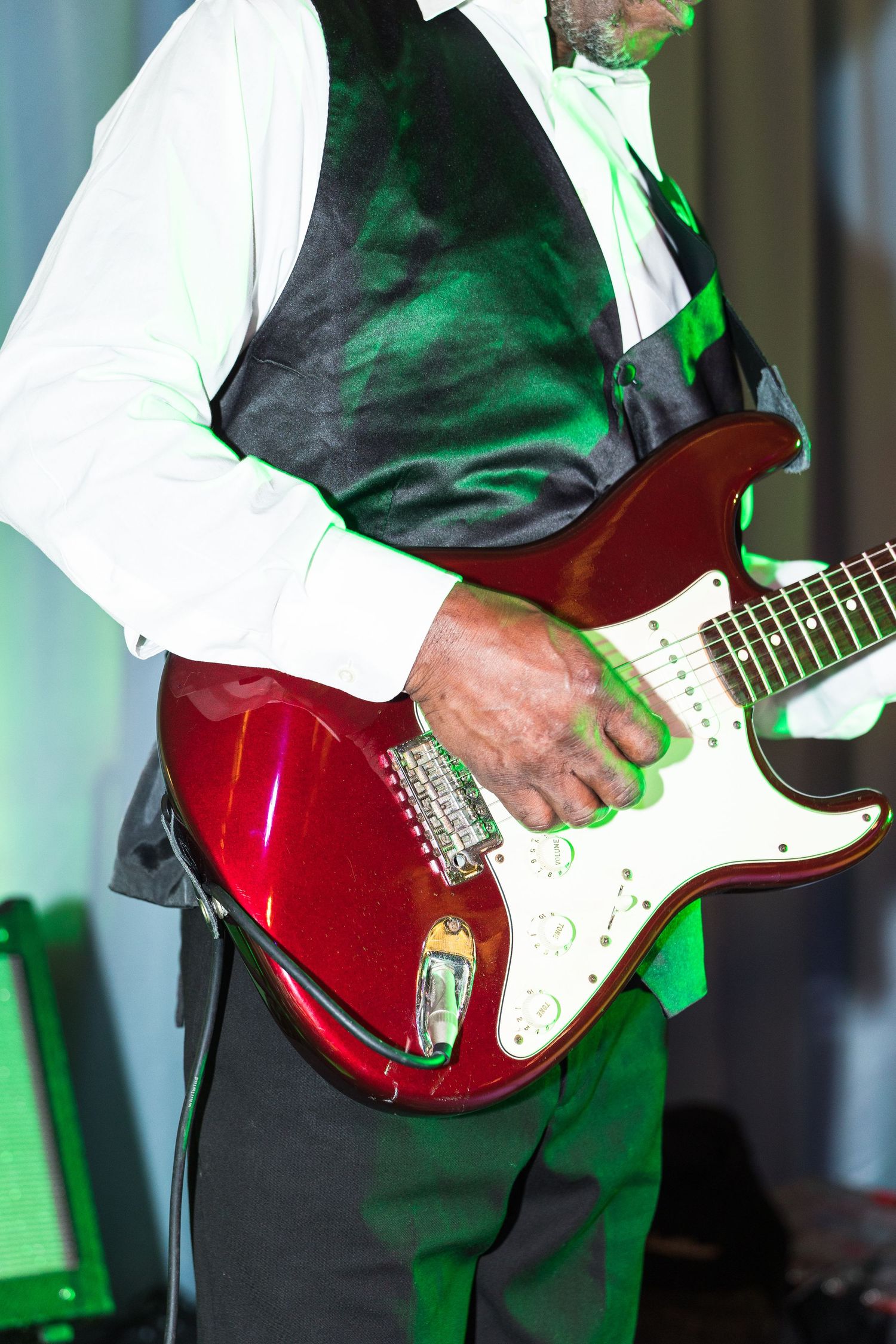 Musician playing a red Stratocaster electric guitar in a white shirt and black vest under green stage lighting.