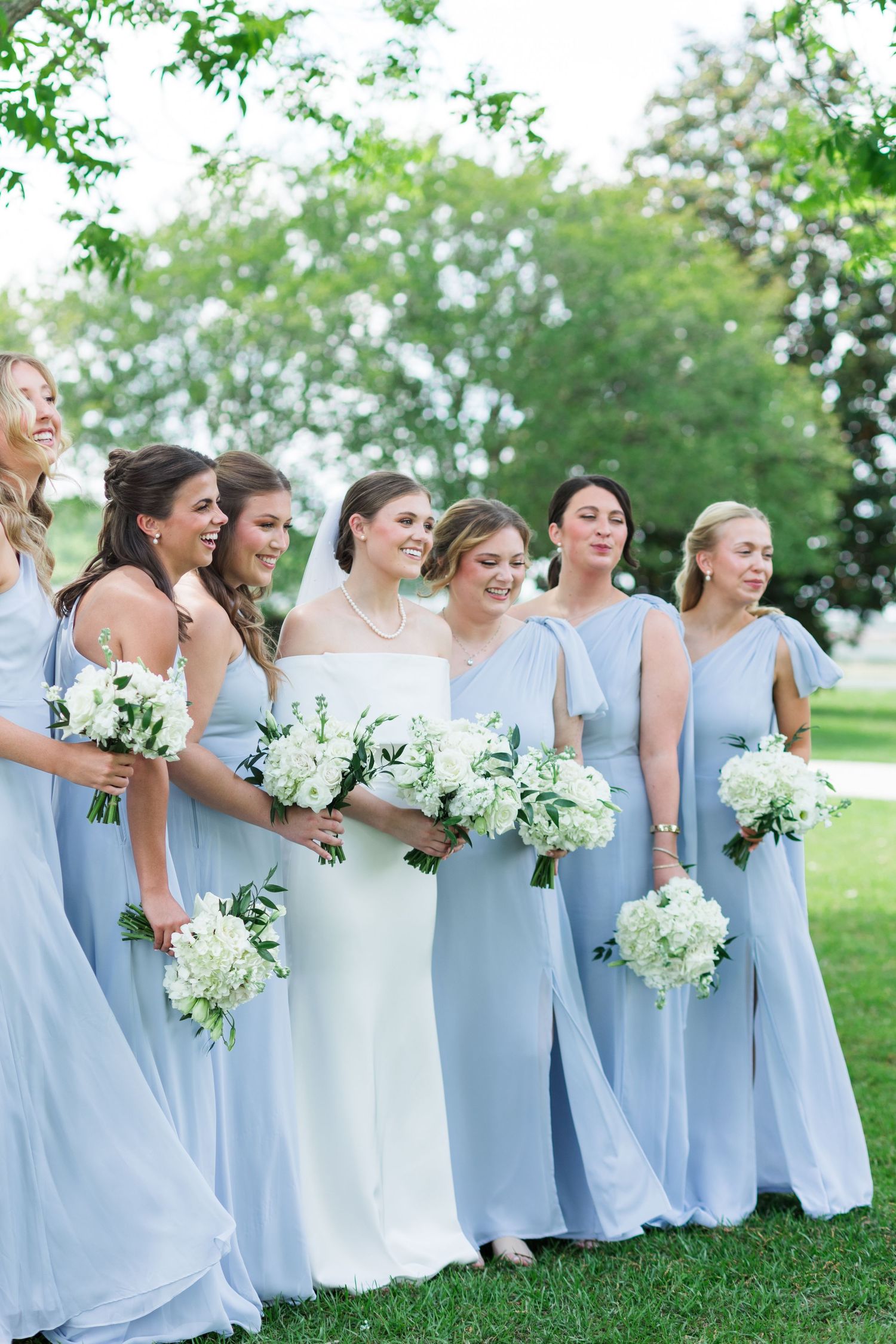Bride in white gown with bridesmaids in light blue dresses holding white floral bouquets outdoors.
