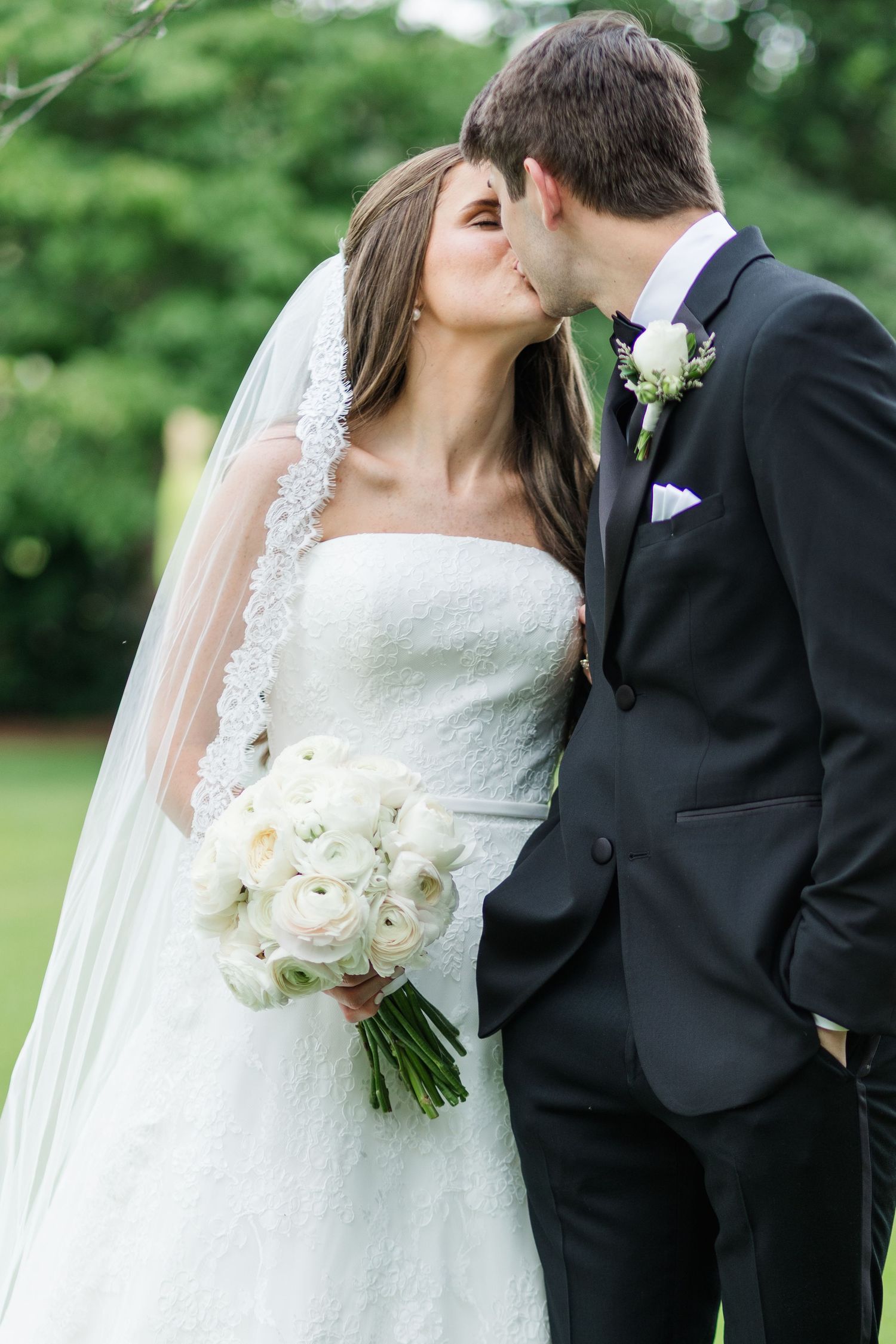 Bride and groom share a kiss outdoors, bride holding white ranunculus bouquet with lace veil.