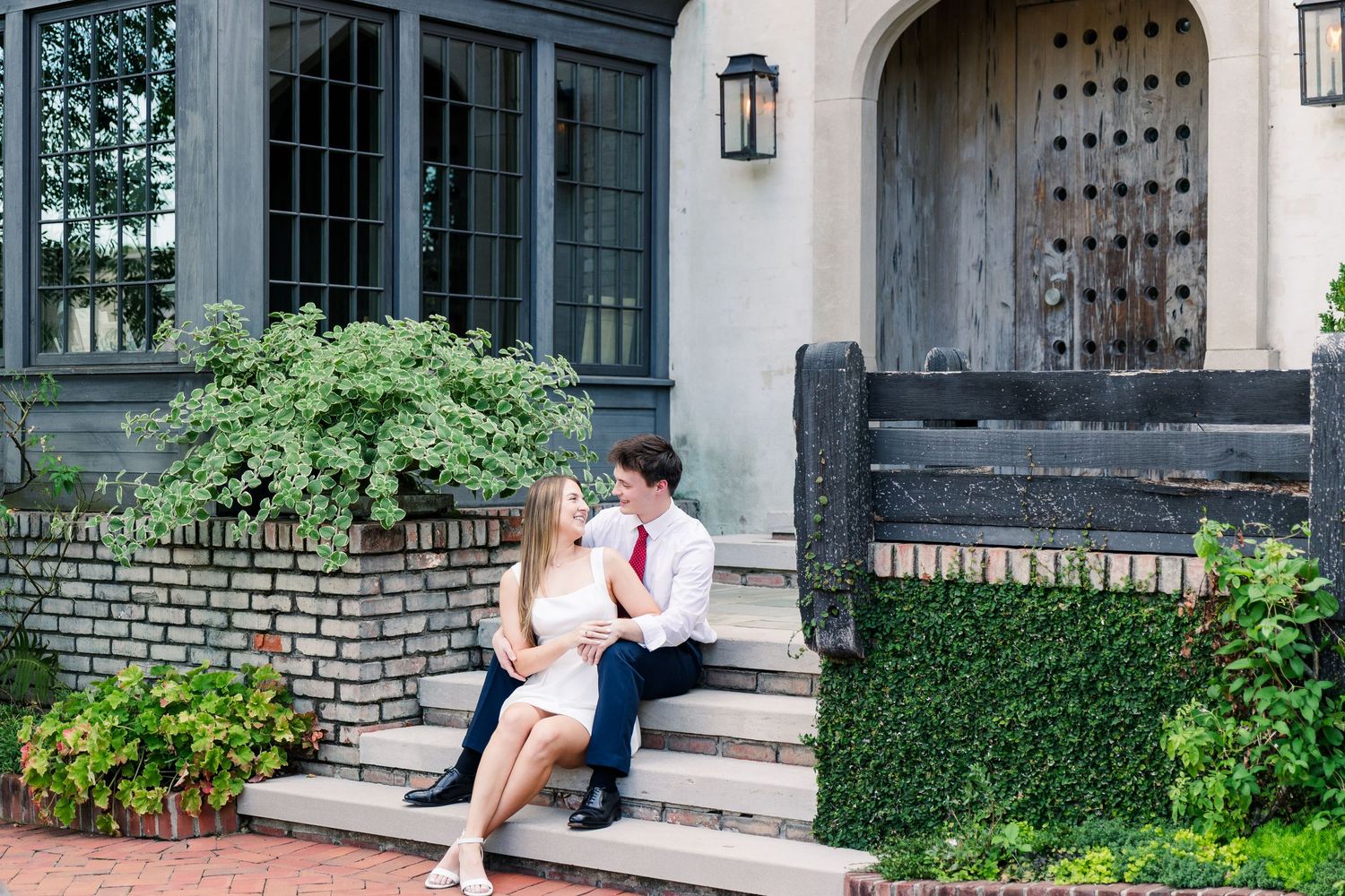 Couple sitting on stone steps of elegant home, sharing a romantic moment surrounded by lush greenery.