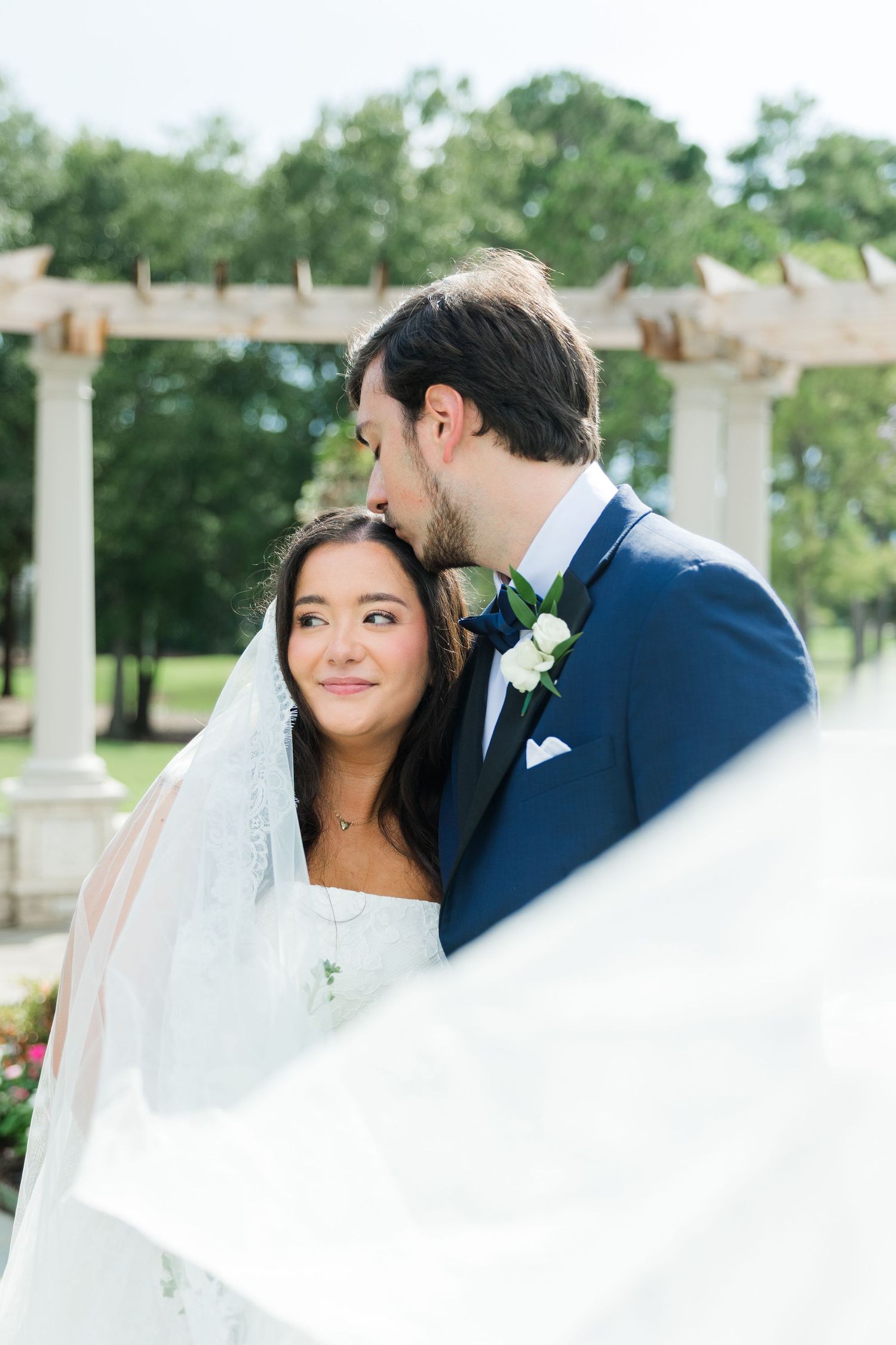 Groom kissing bride on forehead outdoors near pergola, veil flowing in breeze.