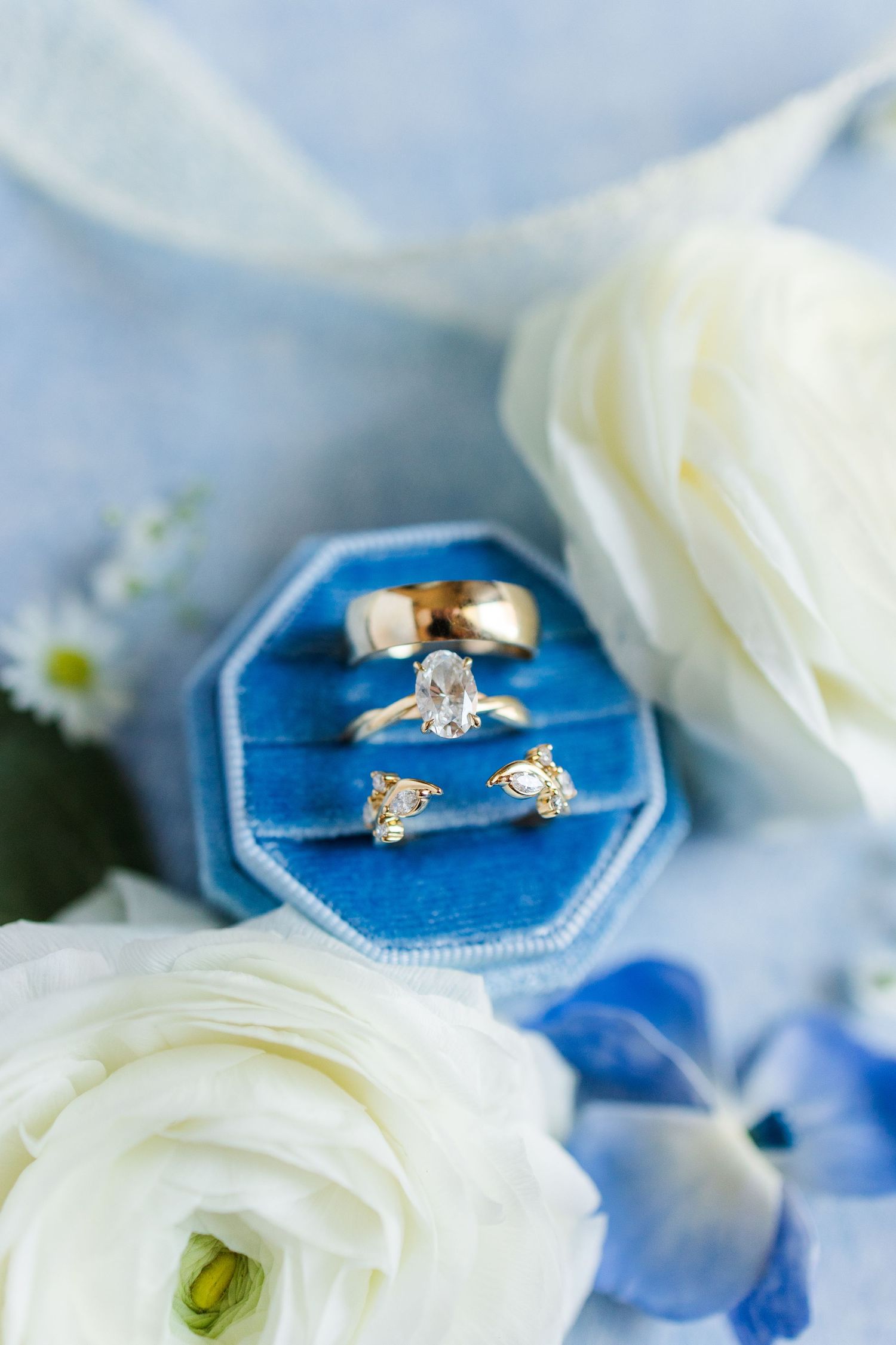 Three gold wedding rings displayed in a blue velvet octagonal ring box surrounded by white ranunculus flowers.