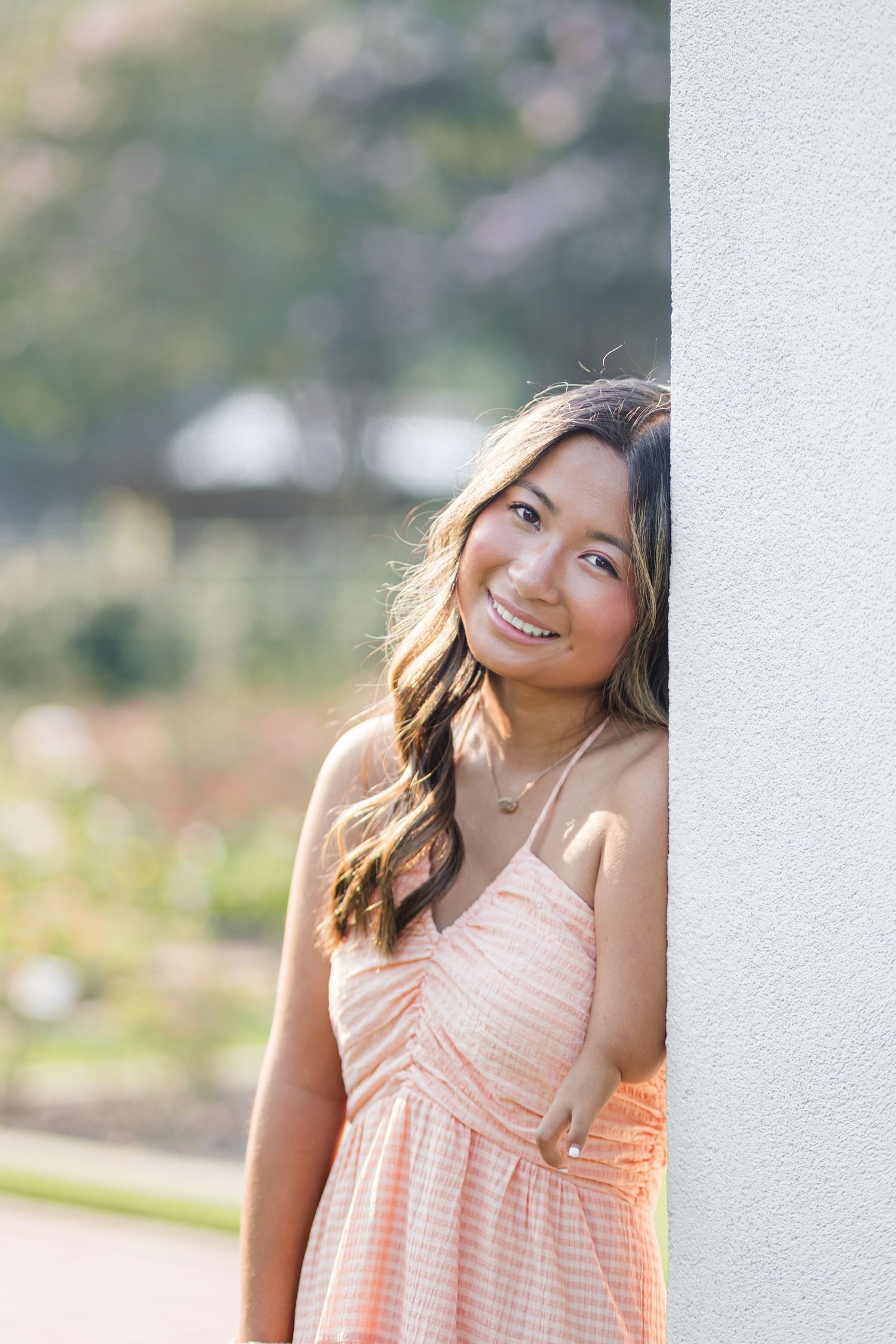 Happy young woman in a peach sundress leaning against a white wall in a sunny garden.