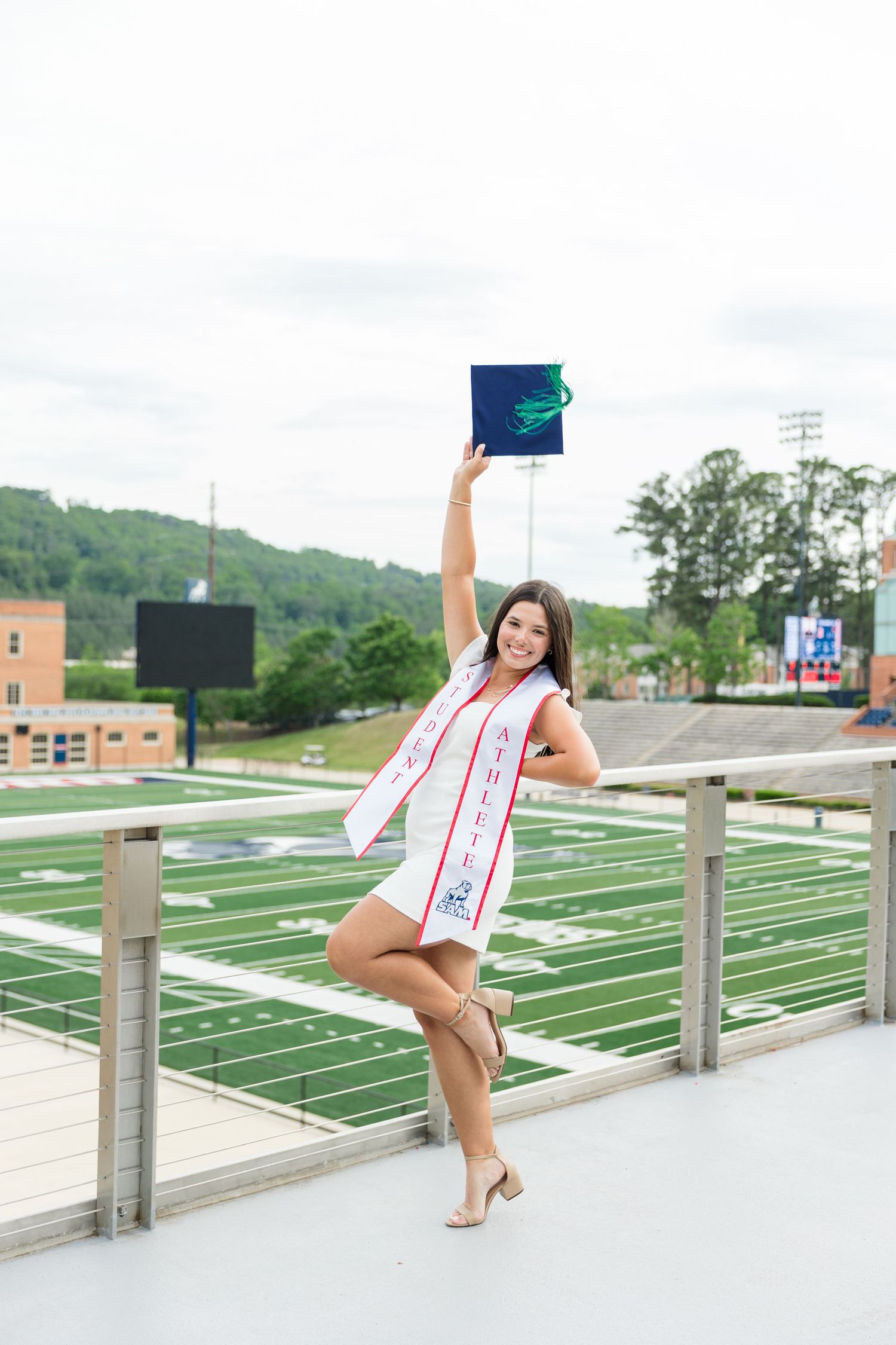 Smiling graduate in white dress and sash raises diploma cap above football stadium backdrop.